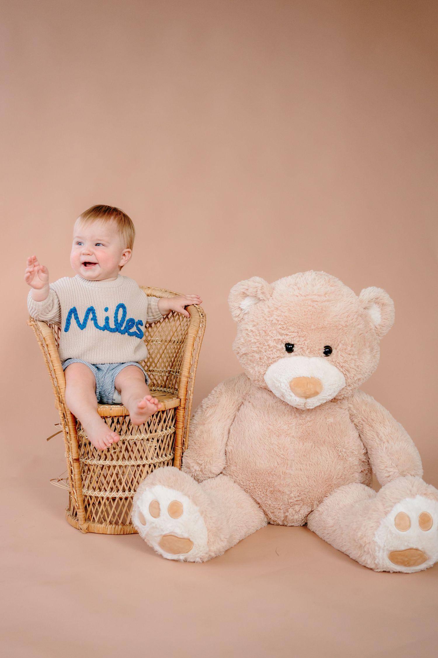 Baby in blue outfit sits in wicker chair next to large plush teddy bear in studio setting.