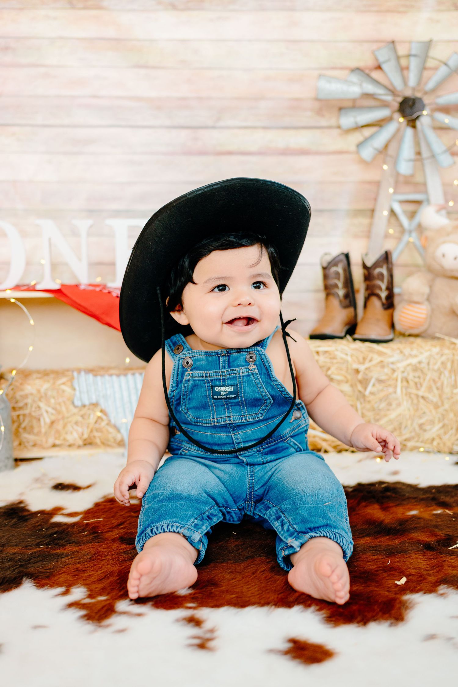 A baby in denim overalls and black cowboy hat sits on a cowhide rug in a rustic western-themed photo shoot setting.