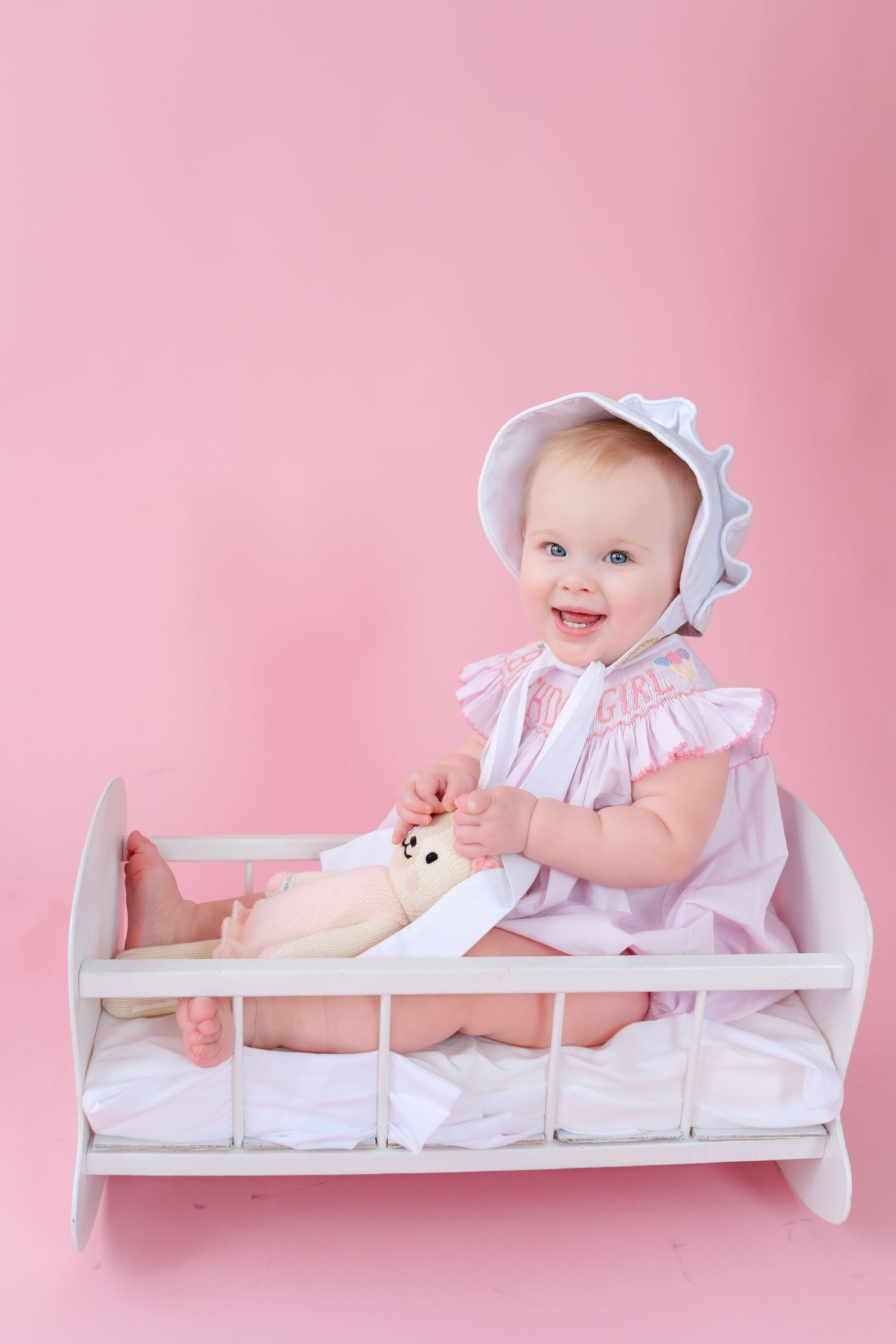 Smiling baby girl in pink dress and bonnet sitting in white toy crib holding stuffed animal on pink background.