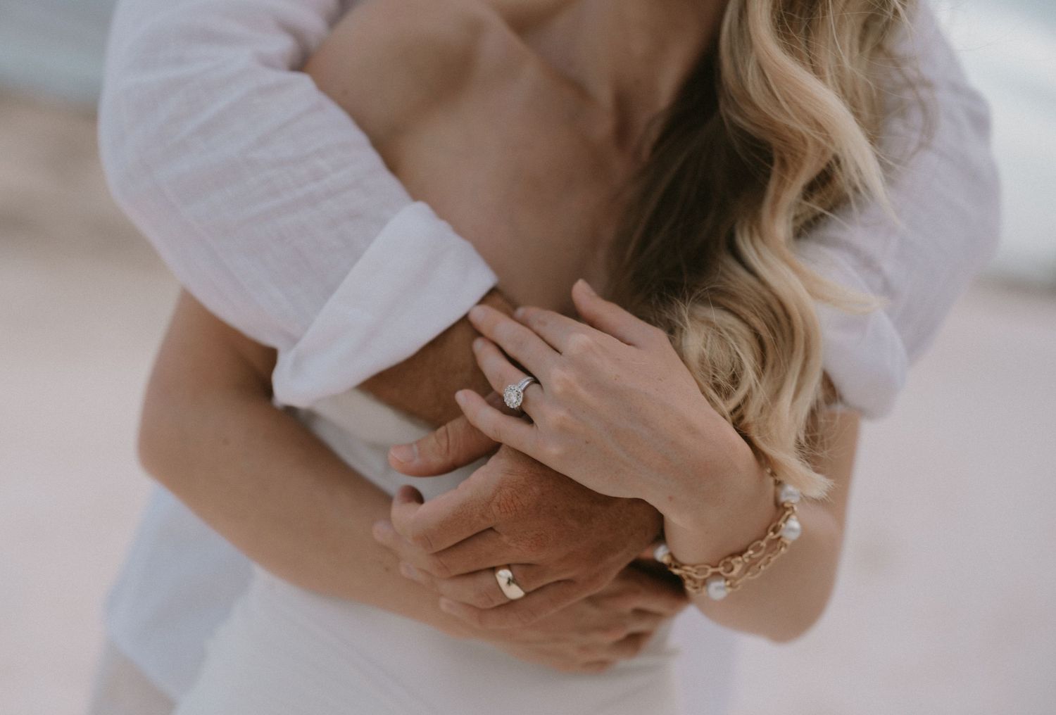 Couple embracing on beach, woman wearing diamond engagement ring and gold bracelet, both dressed in white.