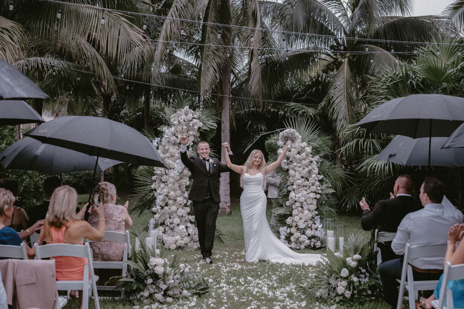 Bride and groom celebrate their recessional under black umbrellas beside a white floral arch and tropical palms, Riviera Maya, Mexico.