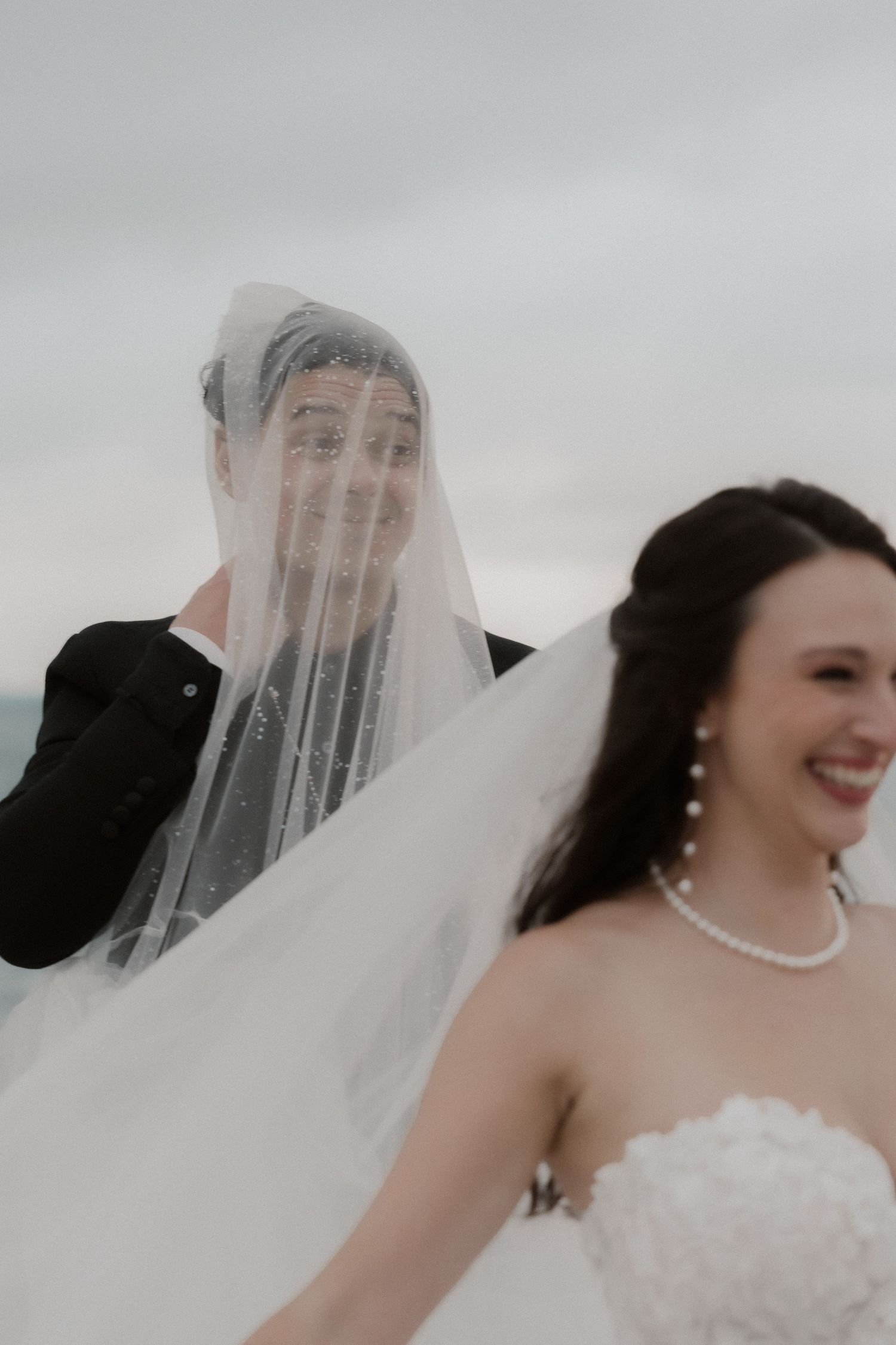 Groom playfully hides under brides veil while she laughs in white gown and pearl necklace outdoors.