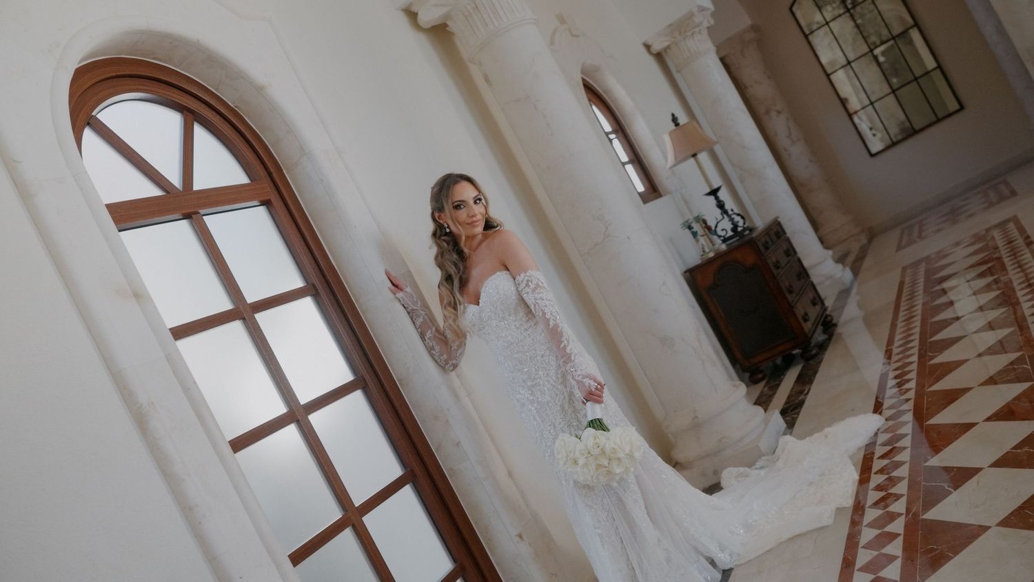 Elegant bride in a white lace gown holding a bouquet, standing by an arched window in a grand venue.