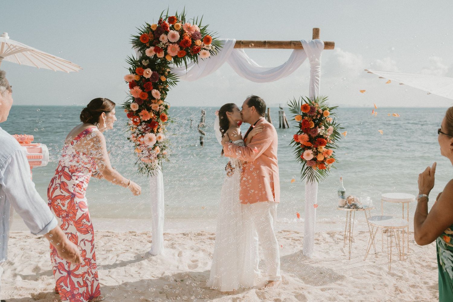 Couple shares a kiss at a beach wedding ceremony beneath a tropical floral arch with the ocean behind them, Playa del Carmen, Mexico.