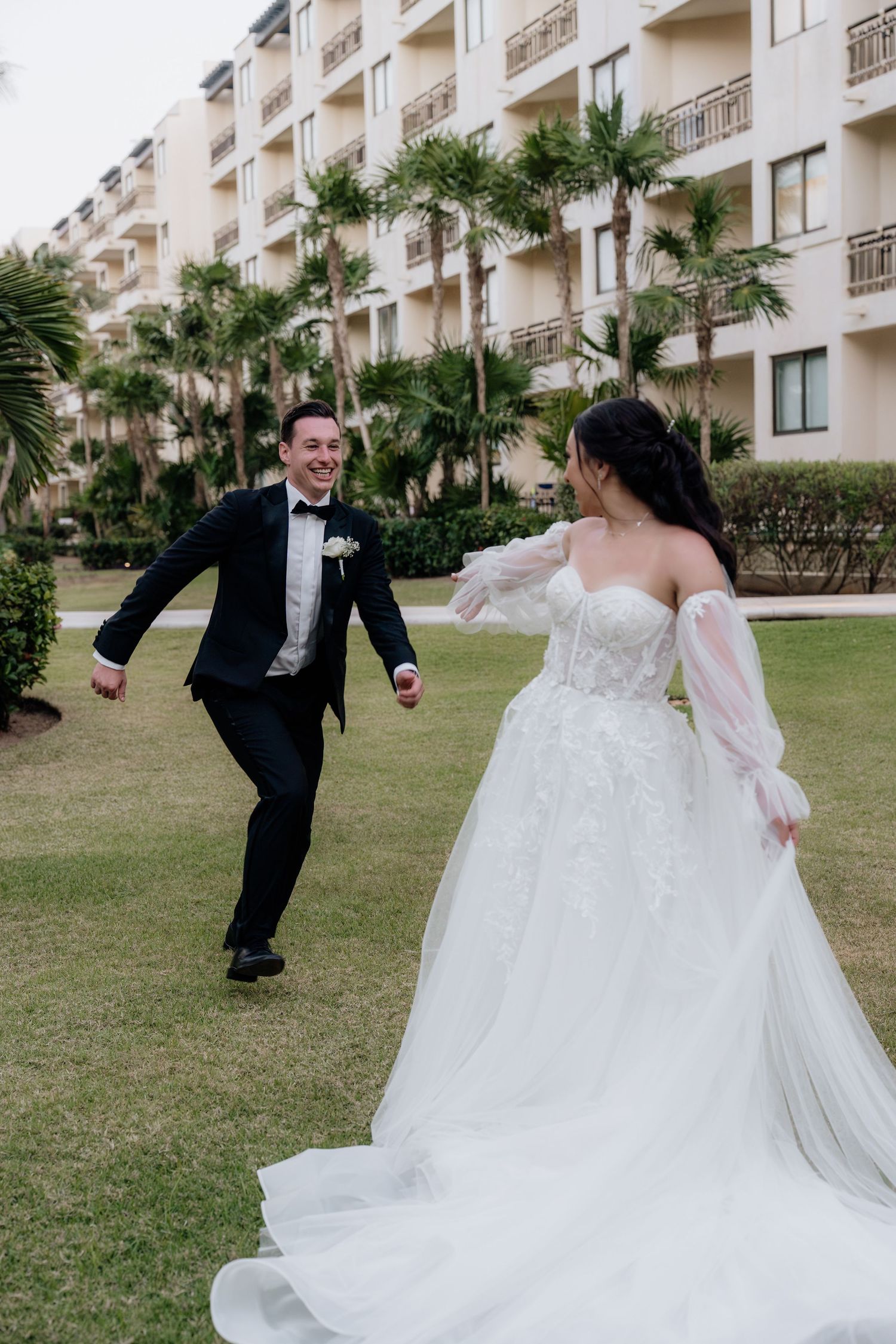 Joyful groom in a tuxedo runs toward his bride in a white ball gown on a resort lawn framed by palm trees, Riviera Maya, Mexico.
