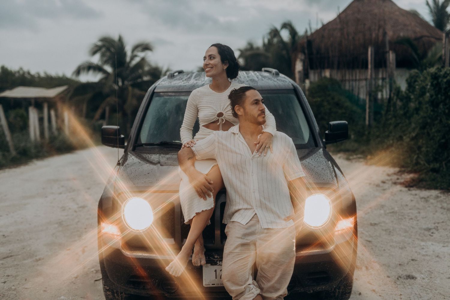Couple posing on car hood at sunset in tropical setting, woman sitting on man's lap, palm trees background.