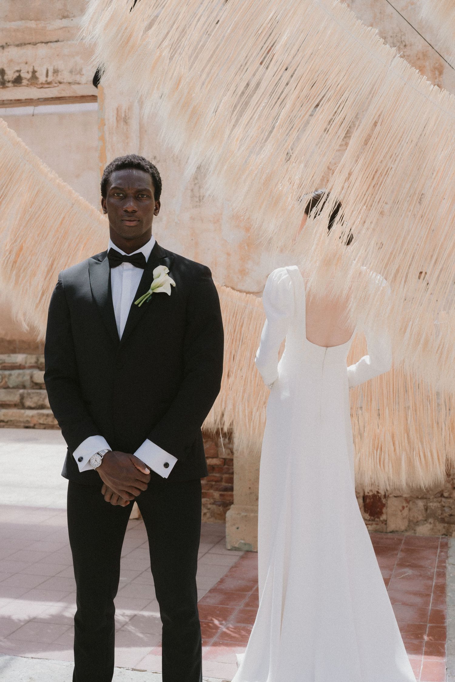 Groom in black tuxedo stands at altar facing bride in white long-sleeve gown during outdoor wedding ceremony.