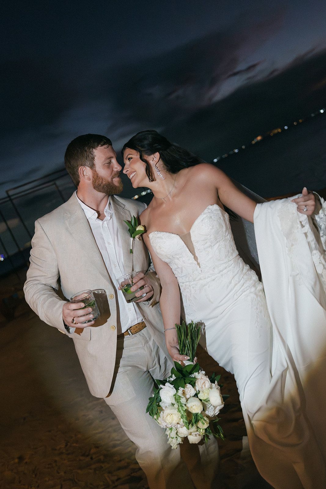Bride and groom share a romantic moment at night, she holds white roses bouquet, he wears tan suit, ocean view behind them.