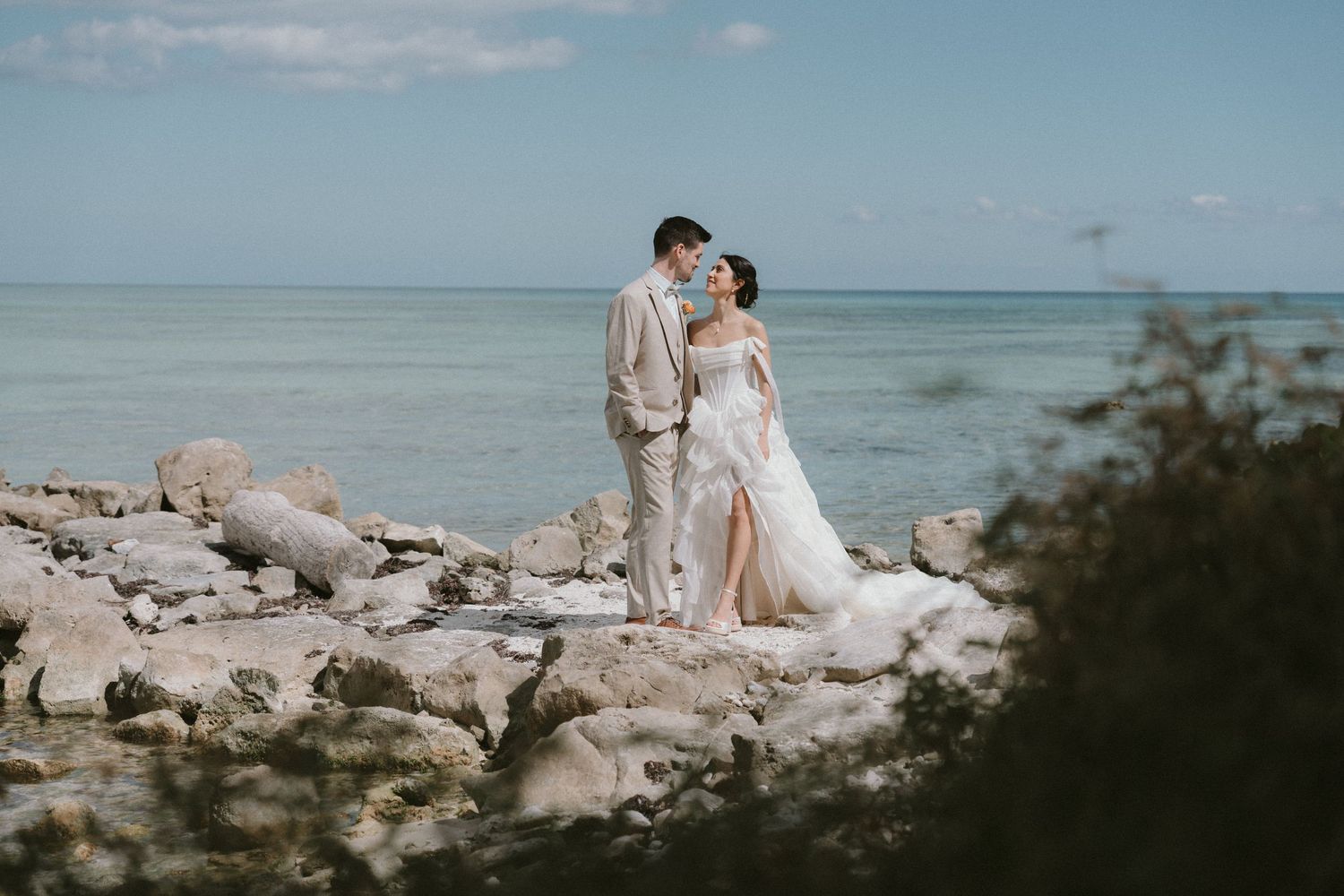 Bride and groom share a quiet moment on a rocky shoreline with a turquoise ocean backdrop, Isla Mujeres, Mexico.