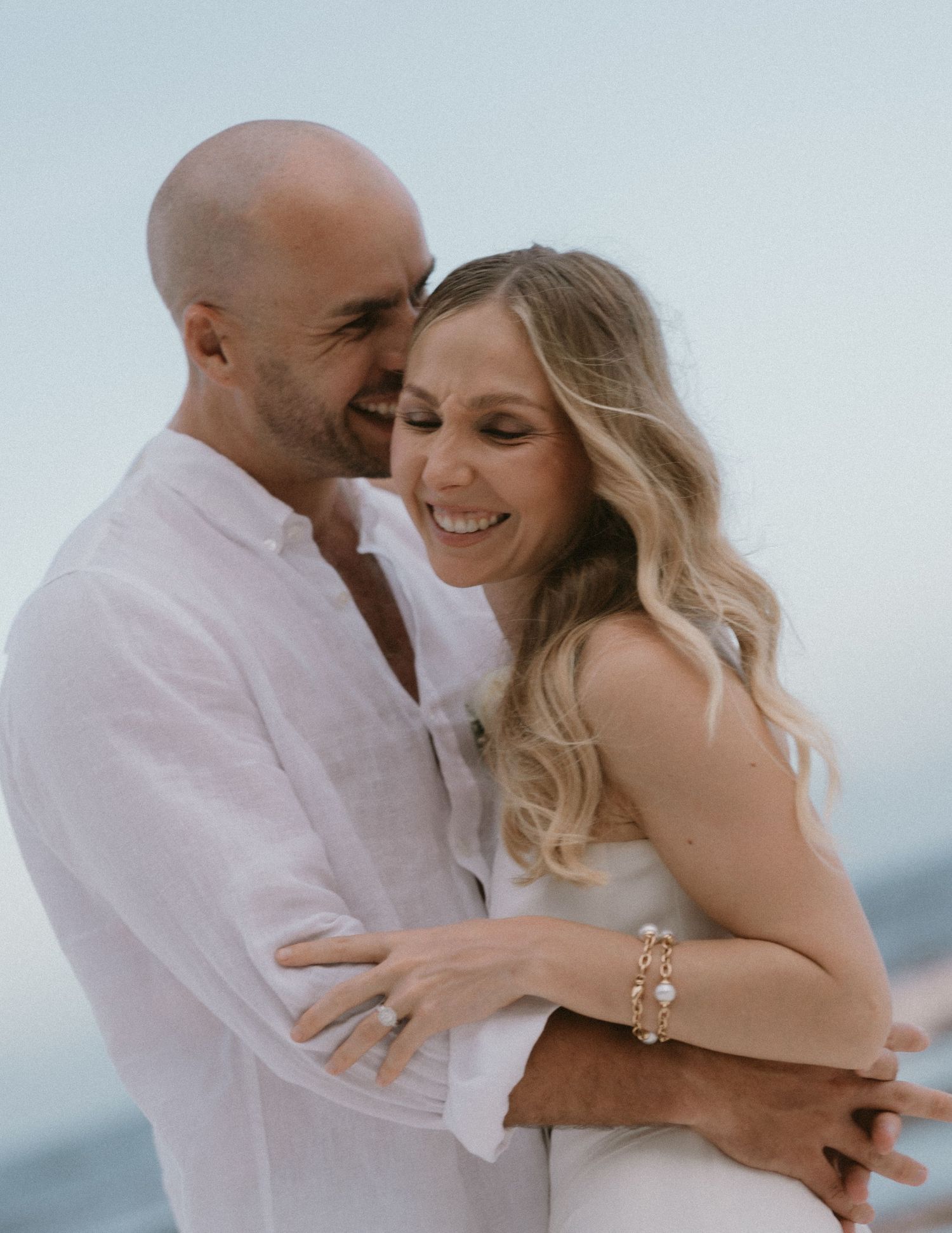 A smiling couple in white attire embraces on a beach, the woman wearing a gold bracelet and engagement ring.