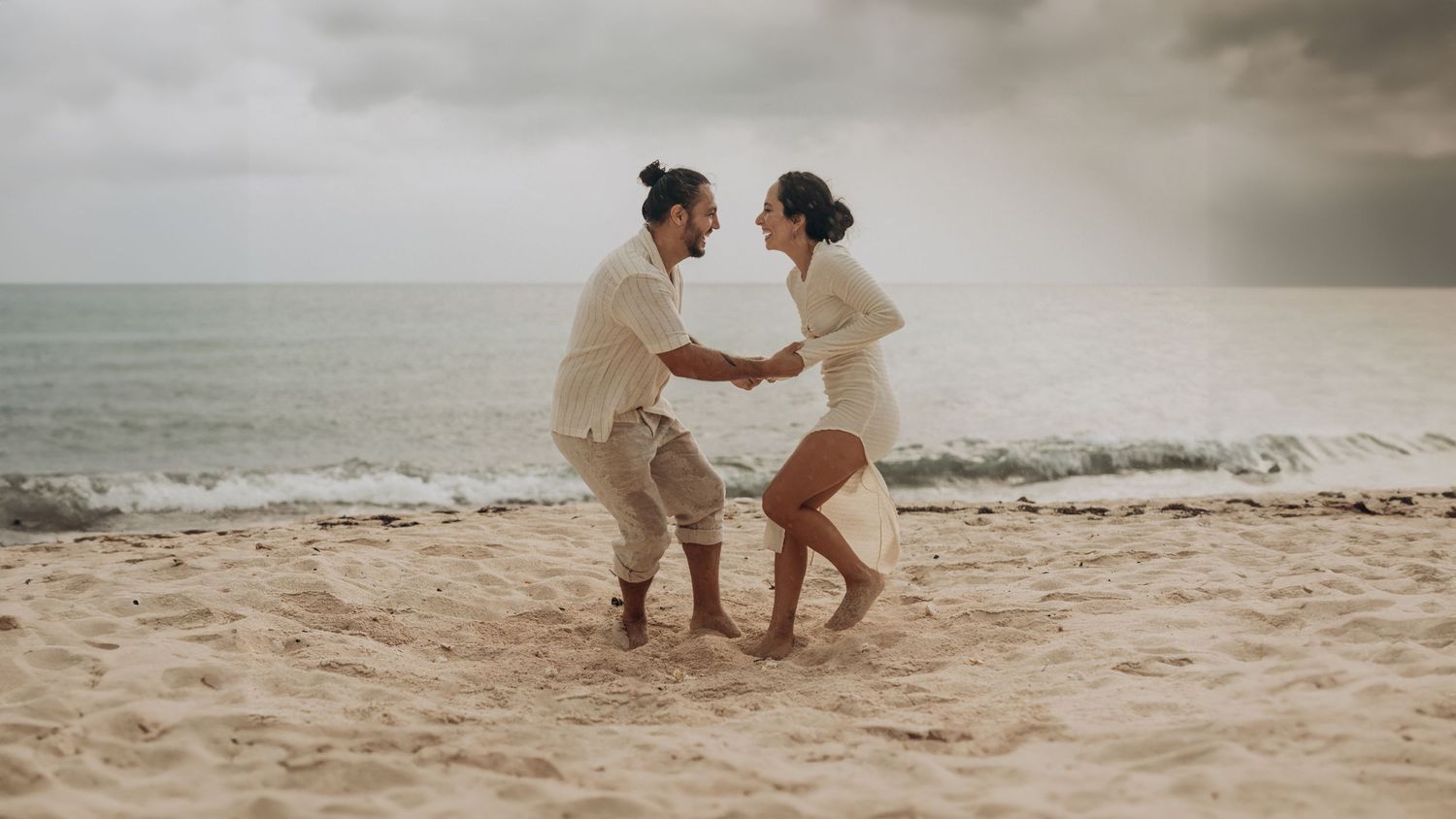 Couple holding hands and dancing playfully on a sandy beach with ocean waves and cloudy sky behind them.