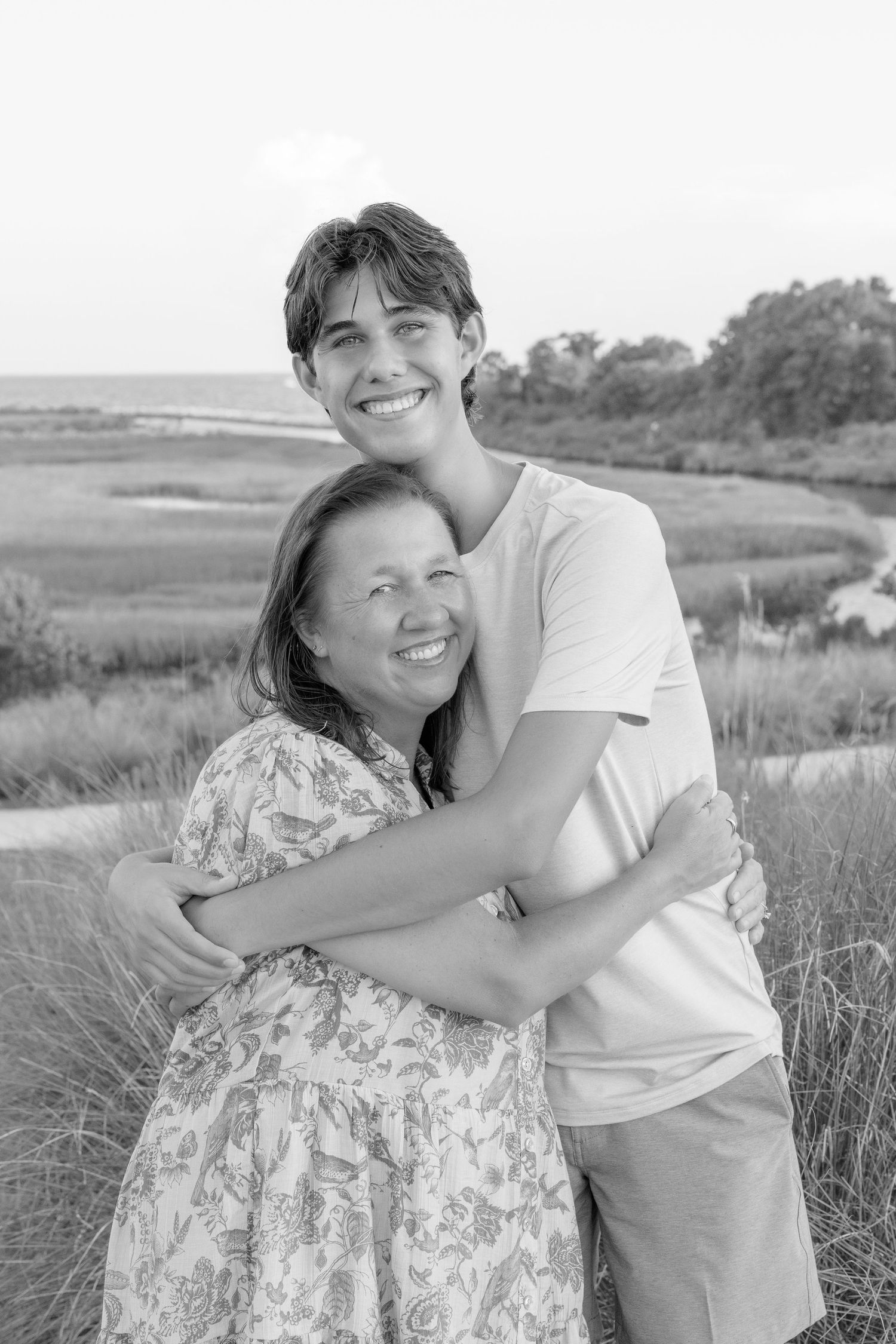 Black and white photo of a young man hugging an older woman outdoors near a coastal marsh, both smiling warmly.