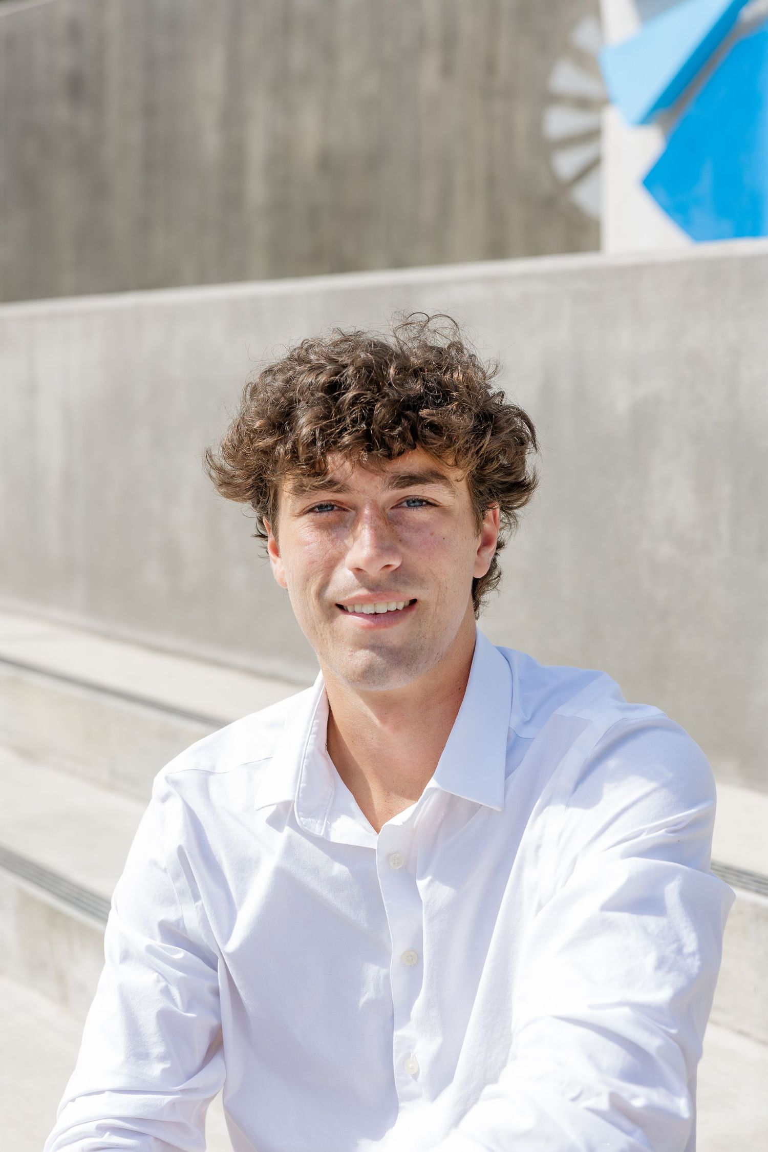 Young man with curly hair in white dress shirt smiling while seated on concrete steps outdoors.