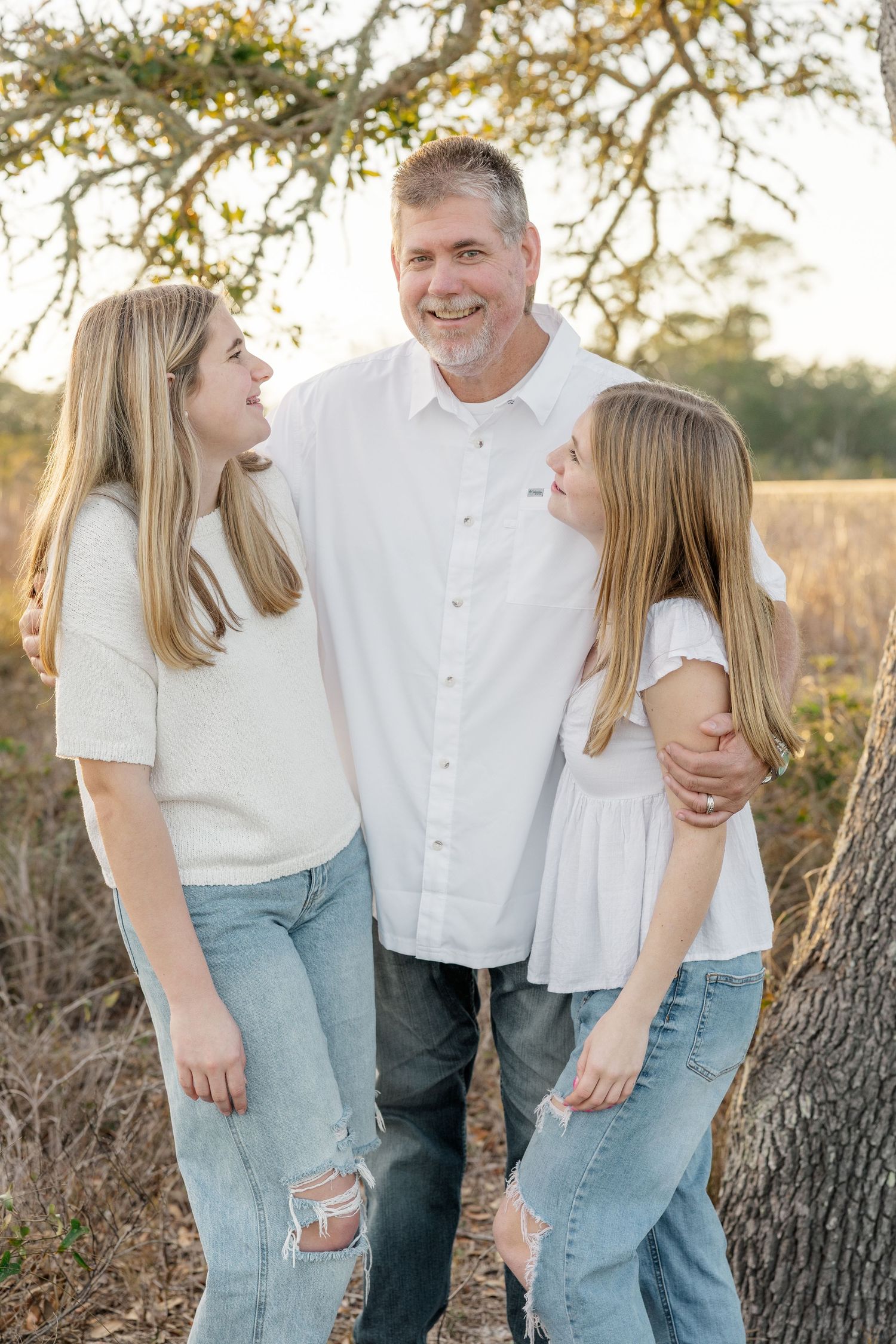 Three people in coordinating white and denim outfits sharing a candid moment together outdoors at sunset.