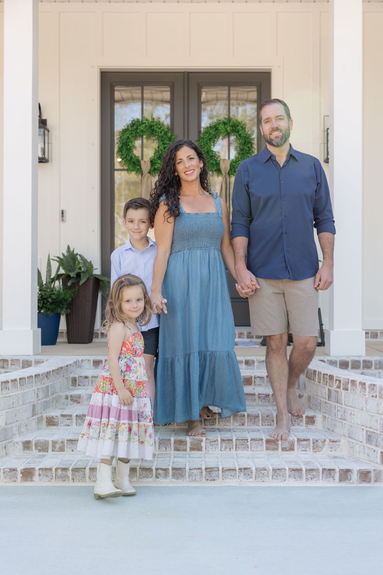Family of four posing on brick front porch steps of white farmhouse home with boxwood wreaths on door.