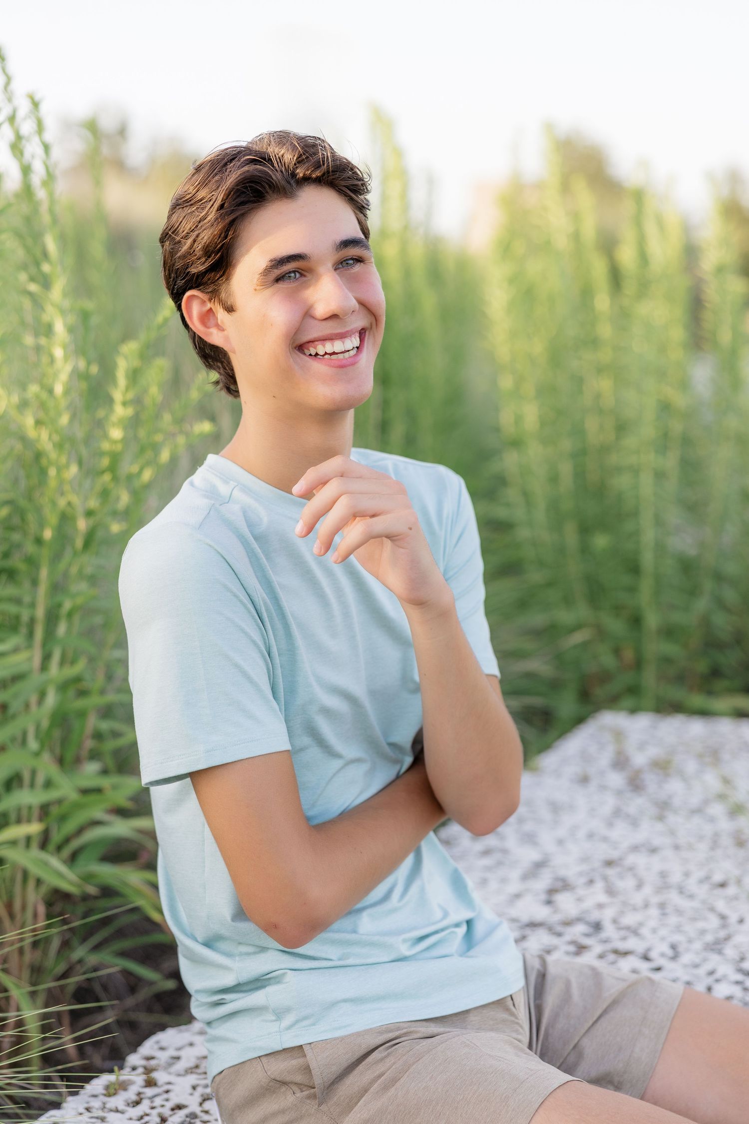 Smiling teenage boy in light blue t-shirt sitting outdoors with green plants in background.