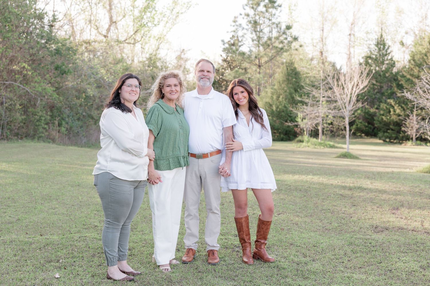 Family of four smiling together outdoors on green grass surrounded by trees on a sunny day.