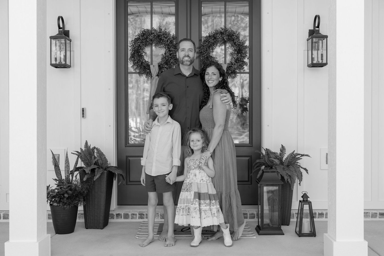 Black and white family portrait of parents with two children posing on a decorated front porch with heart wreaths.