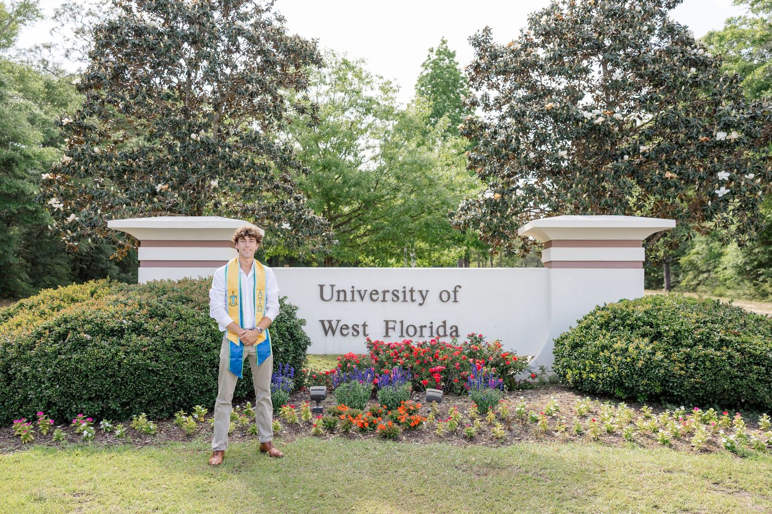 Graduate poses proudly in front of the University of West Florida sign surrounded by colorful flowers and lush greenery.
