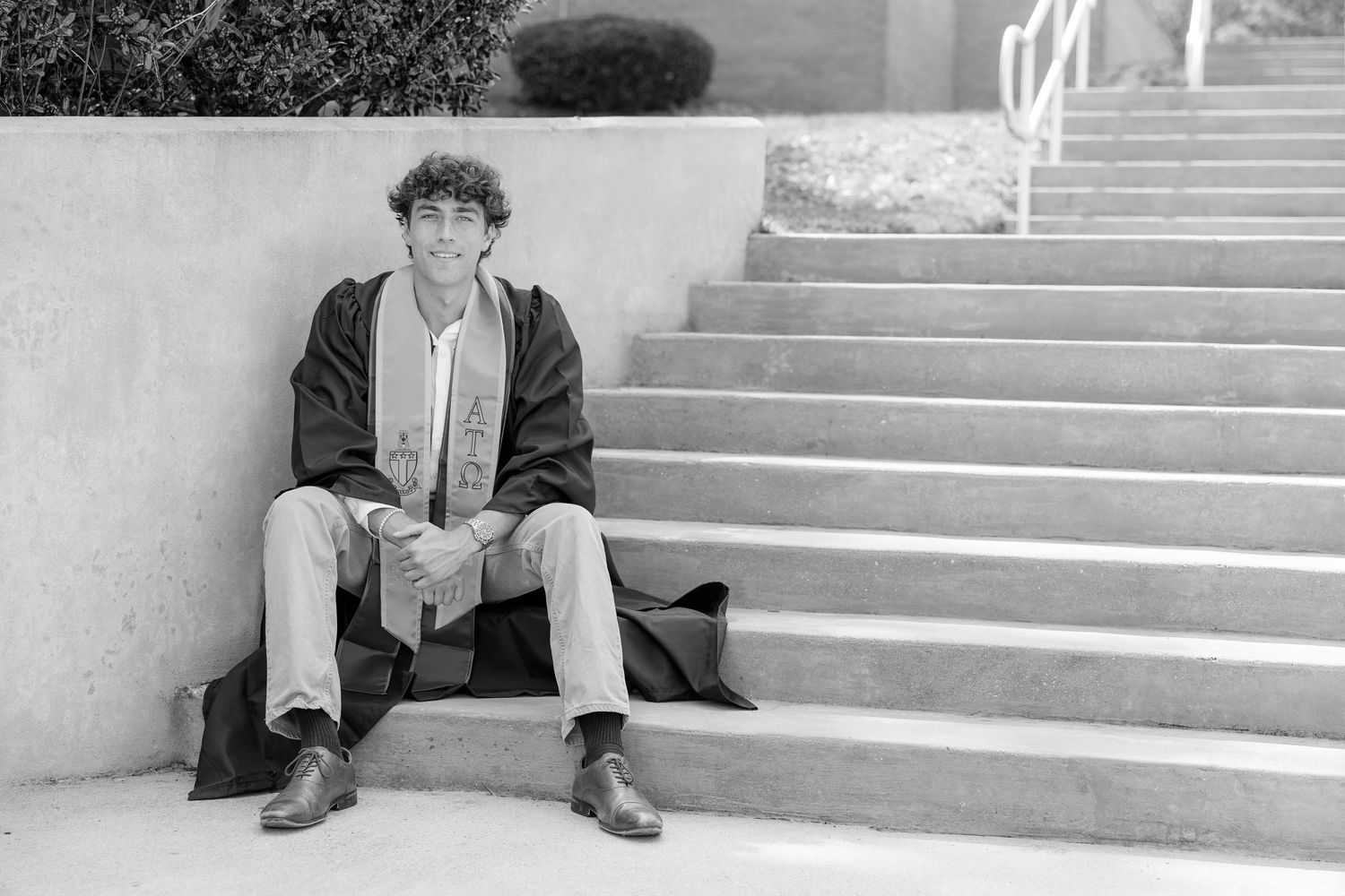 Black and white photo of a young man sitting on concrete steps outdoors, wearing a long coat and casual pants.