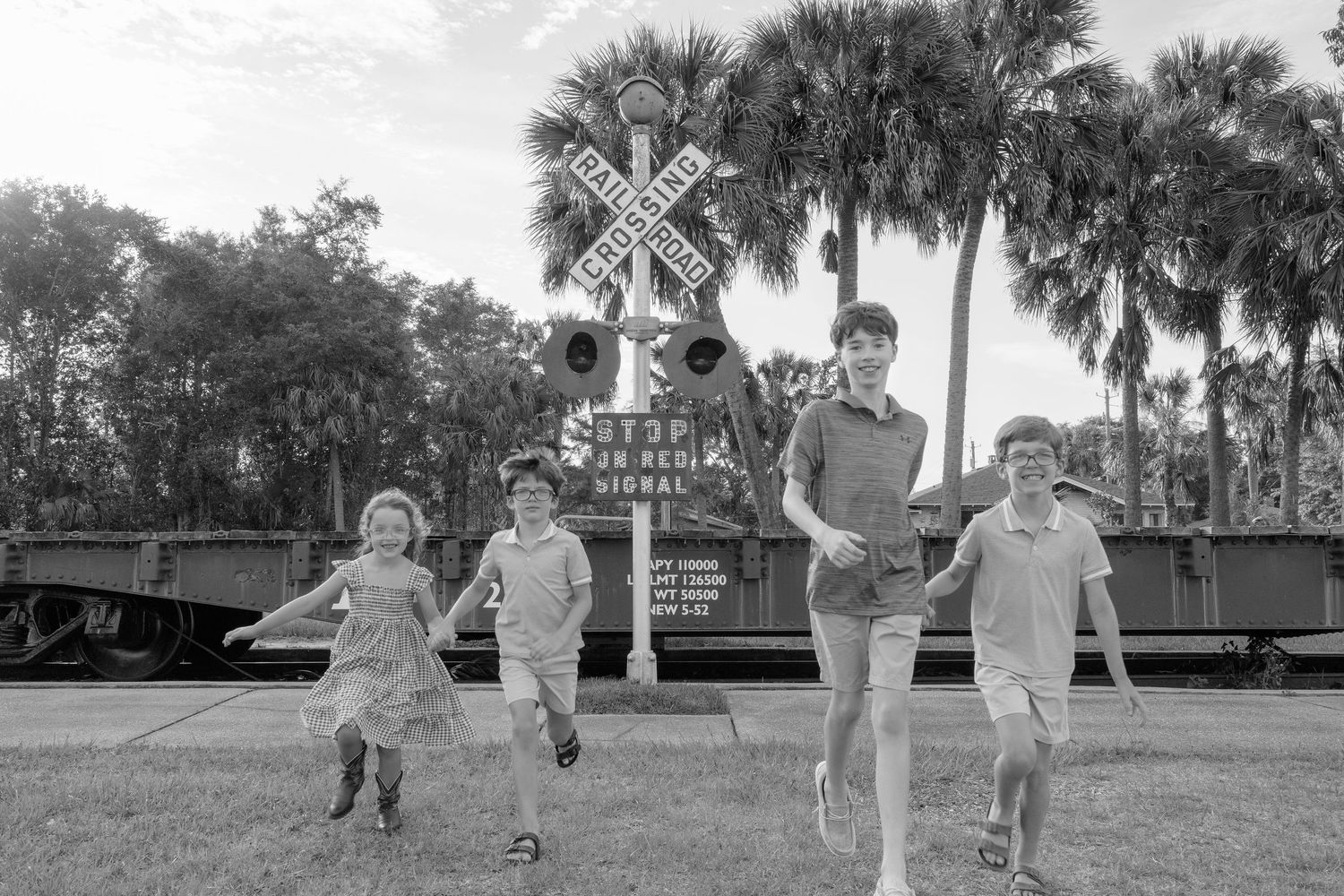 Four children run joyfully near a railroad crossing sign with palm trees and a train car in a black and white photo.