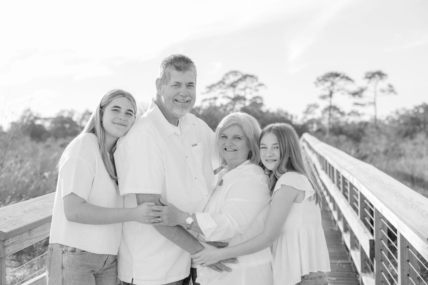 A family group poses together on a wooden boardwalk with pine trees visible in the background.