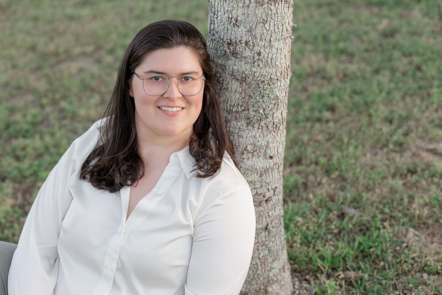 Woman with glasses and dark hair smiles while leaning against a tree outdoors in a white blouse.