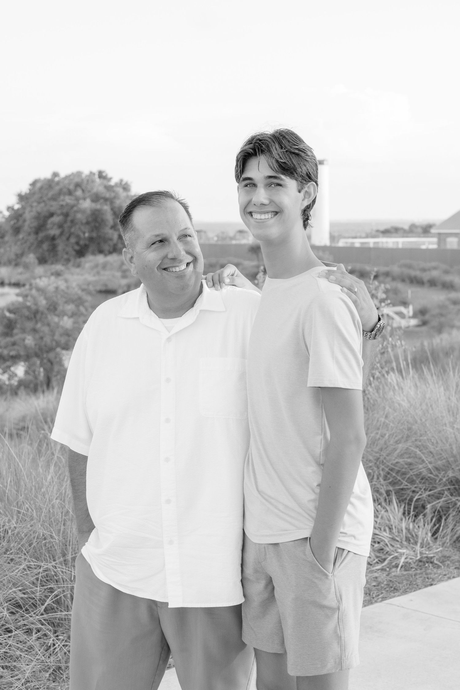 Father and teenage son smiling together outdoors in black and white photo, wearing casual shirts and shorts.