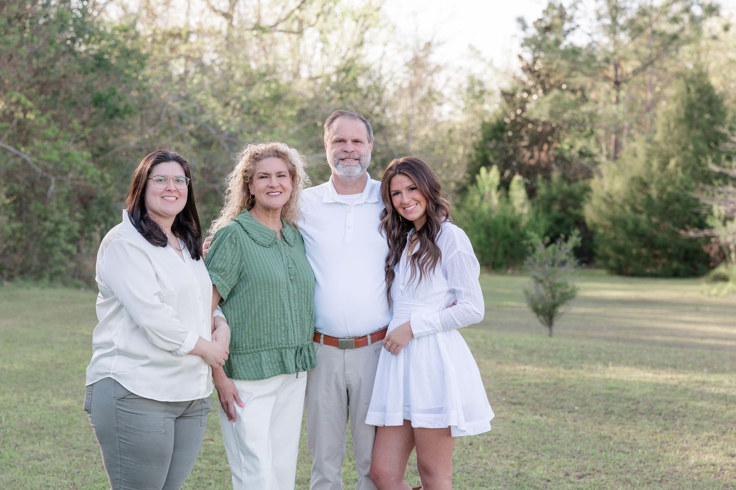 Family of four smiling together outdoors on green lawn with trees in background, wearing white and green outfits.