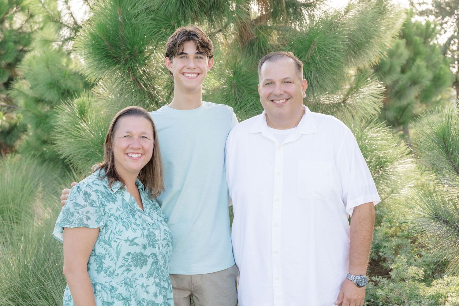 Family of three smiling outdoors among pine trees, wearing light casual clothing in a warm summer setting.