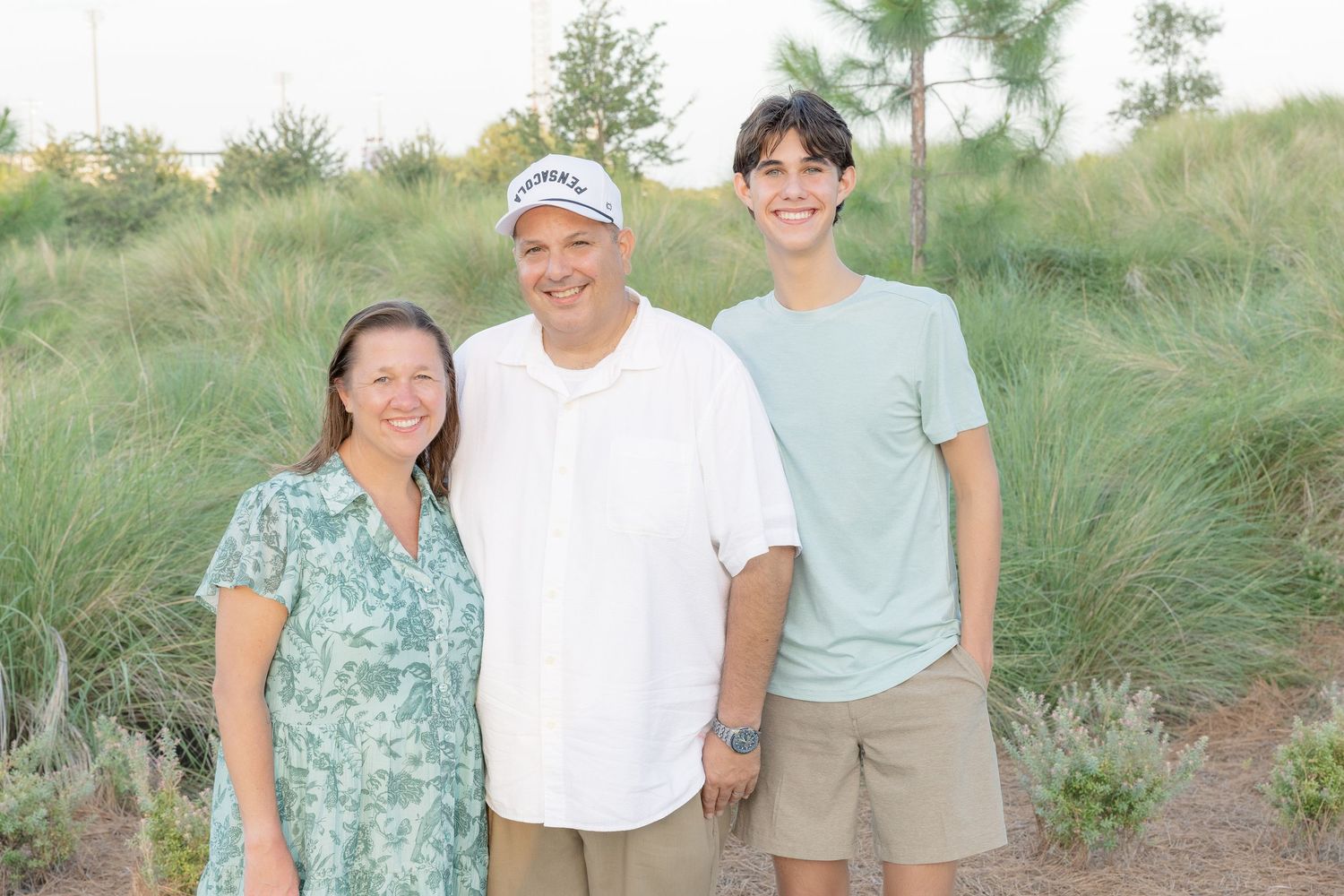 Family of three smiling together outdoors surrounded by lush green bushes in warm evening light.