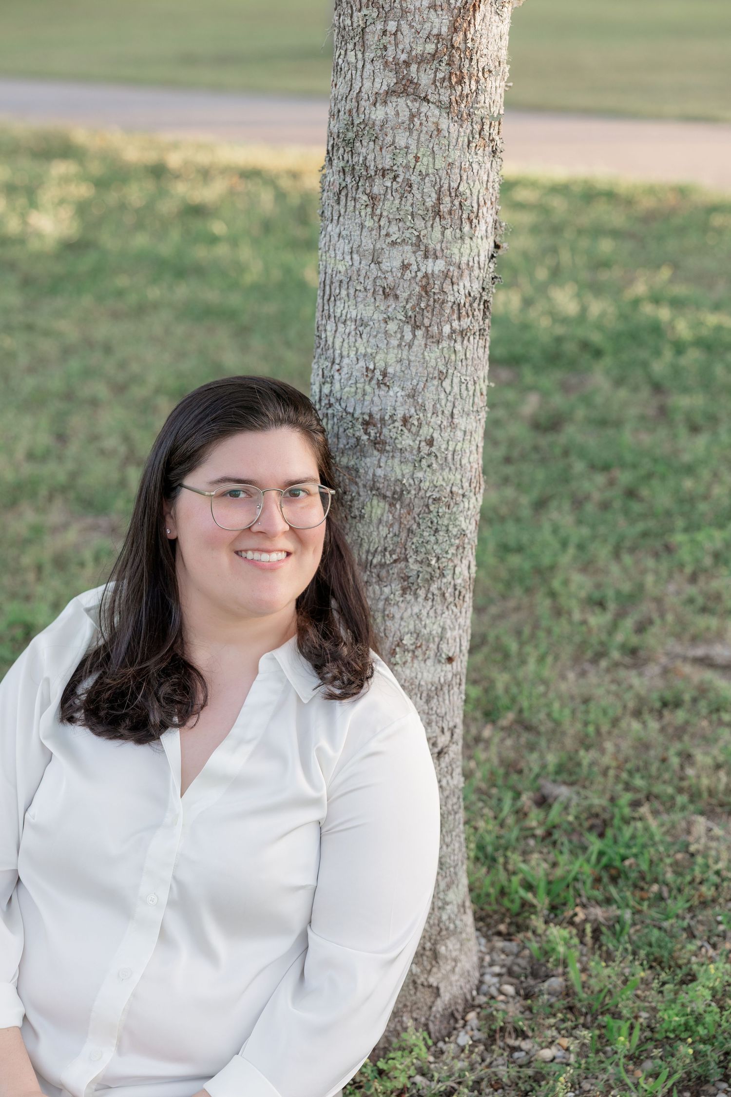 Smiling woman with glasses in white blouse leaning against tree trunk in green park setting.