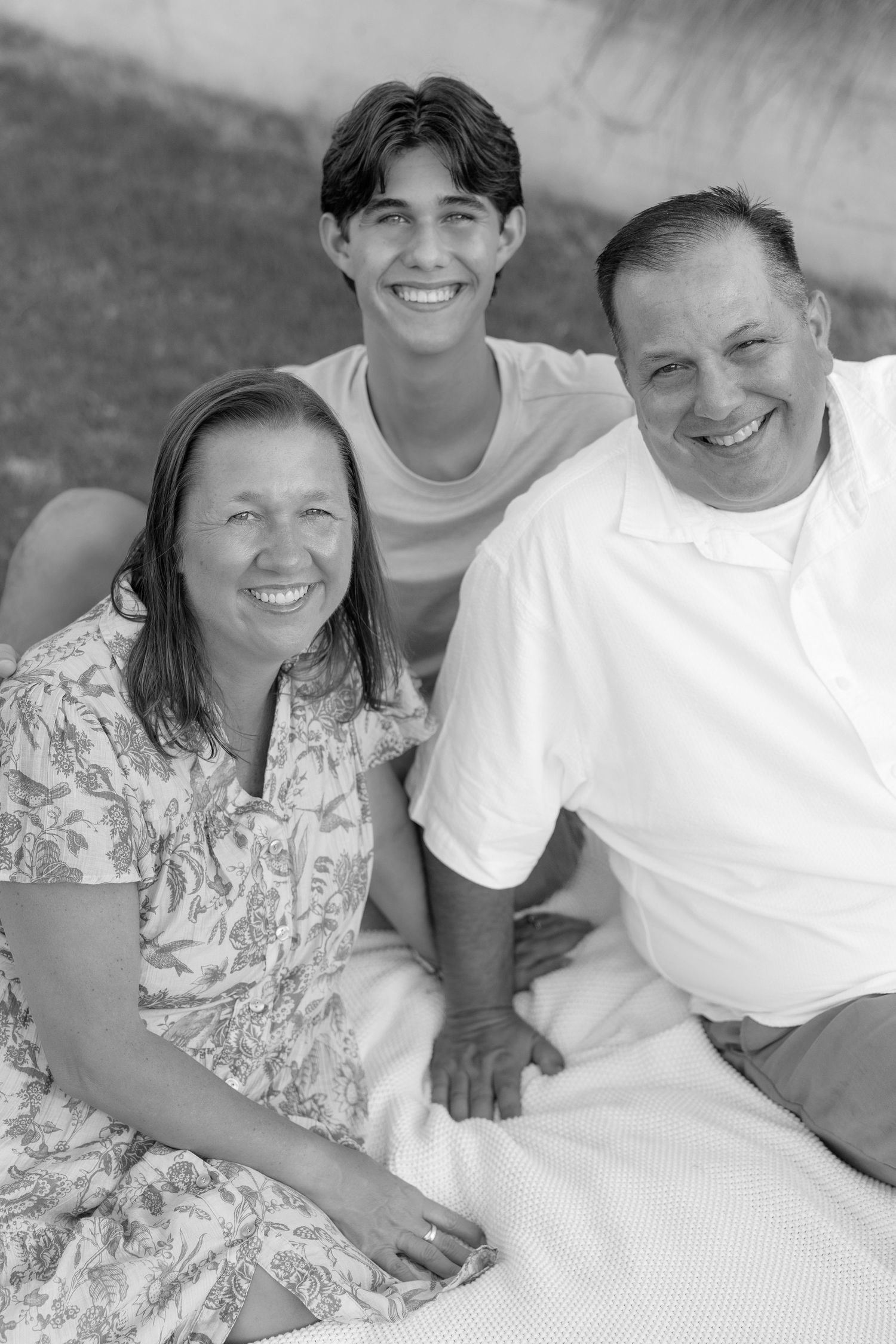 Black and white family portrait of smiling parents and teenage son posing together outdoors.
