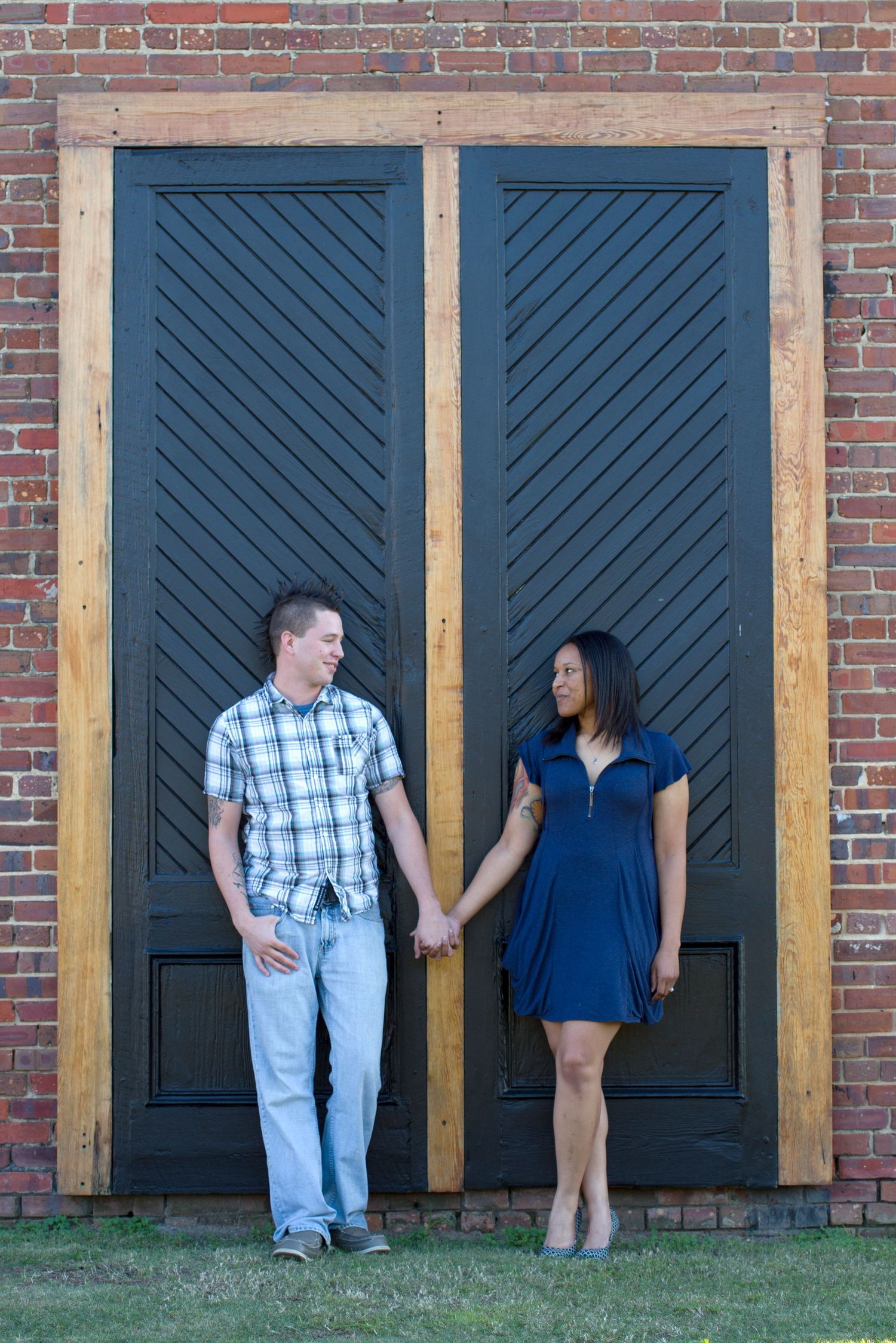 A couple holds hands while standing against large blue chevron-patterned wooden doors on a brick wall.