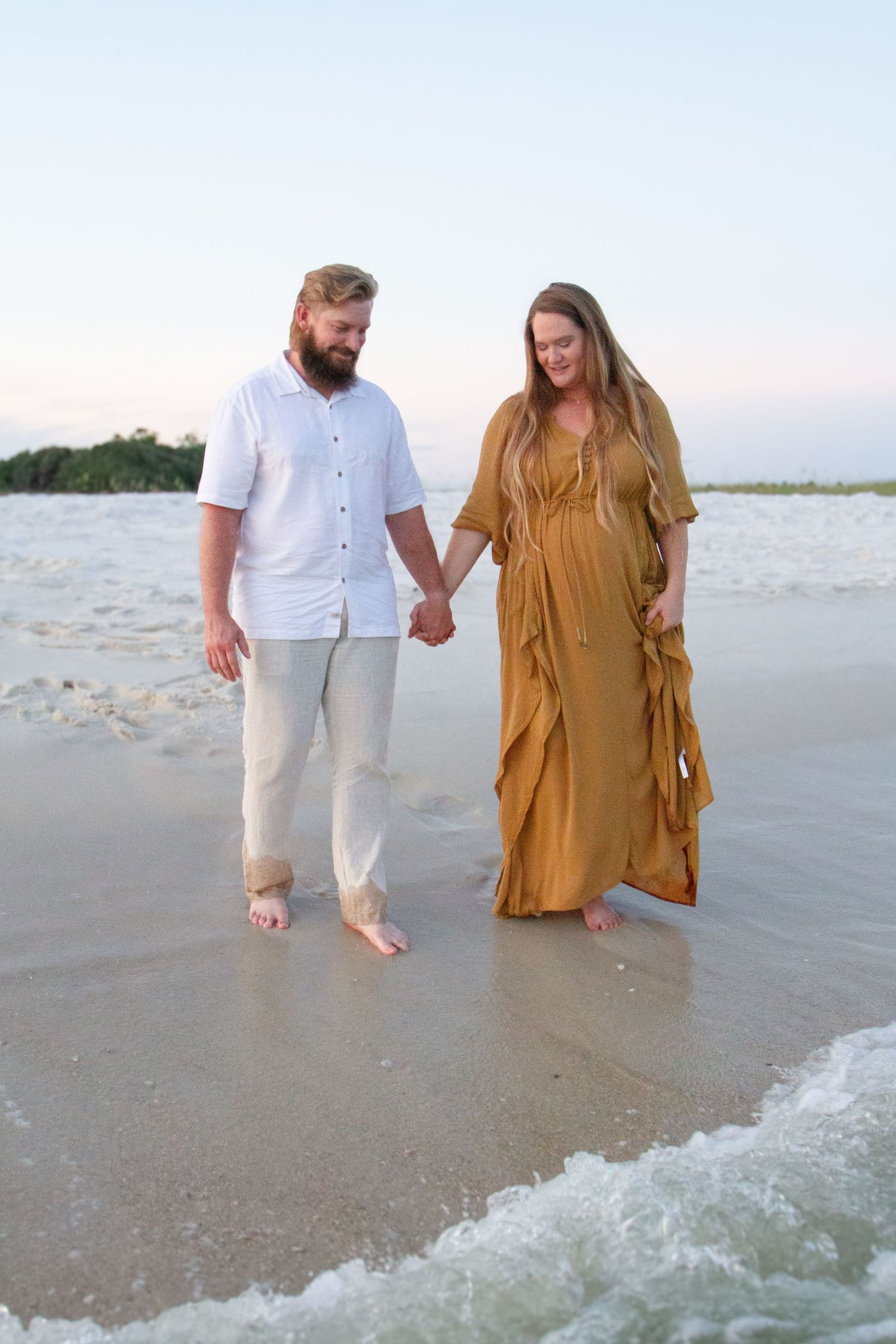 A couple walks hand in hand on a sandy beach as waves roll in during golden hour.