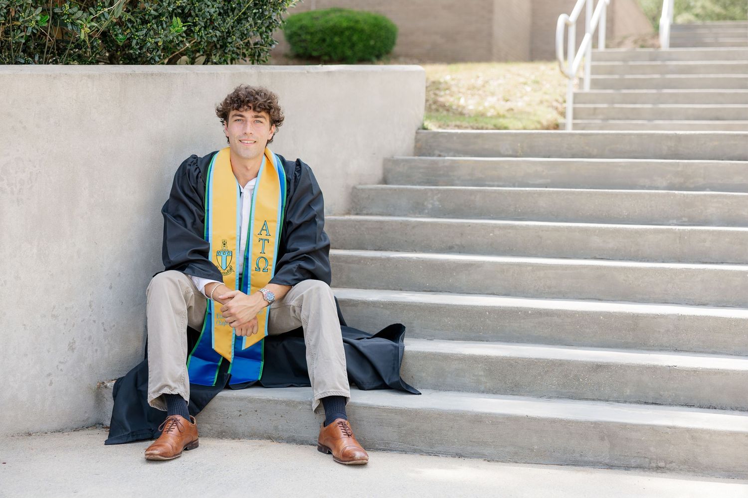 Young man in graduation gown and colorful stole sitting on concrete steps, smiling for senior portrait photo.