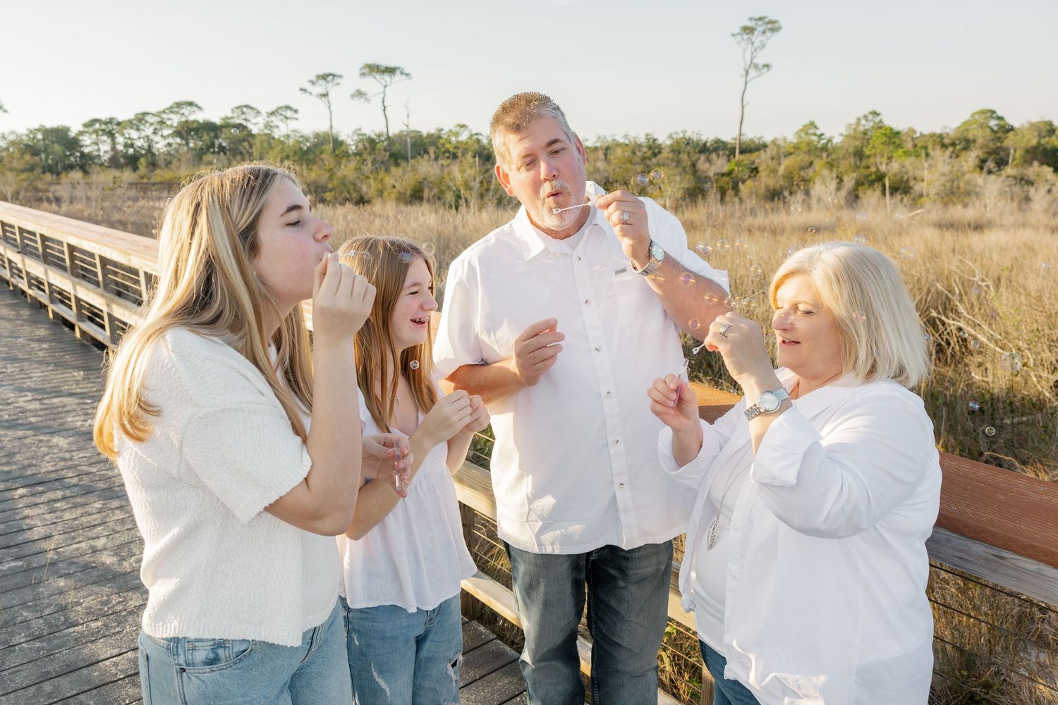 Family wearing coordinated white outfits poses playfully together on a beach boardwalk at sunset.