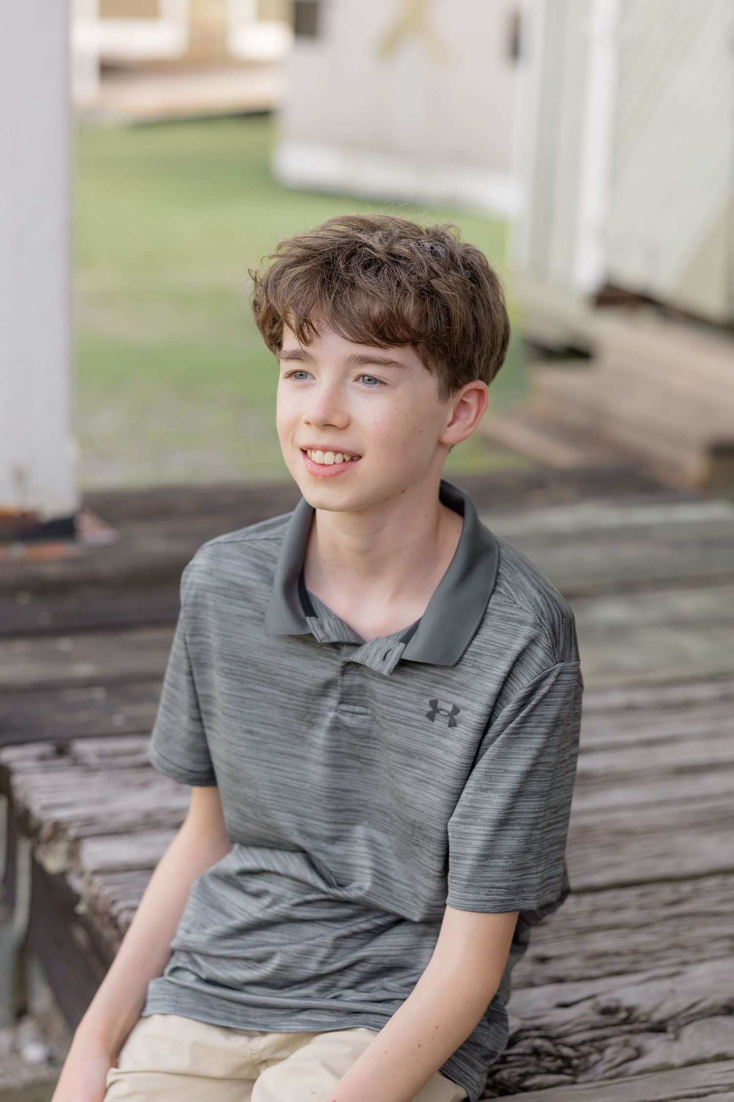 Smiling young boy with brown hair wearing a gray Under Armour polo shirt, sitting on wooden steps outdoors.