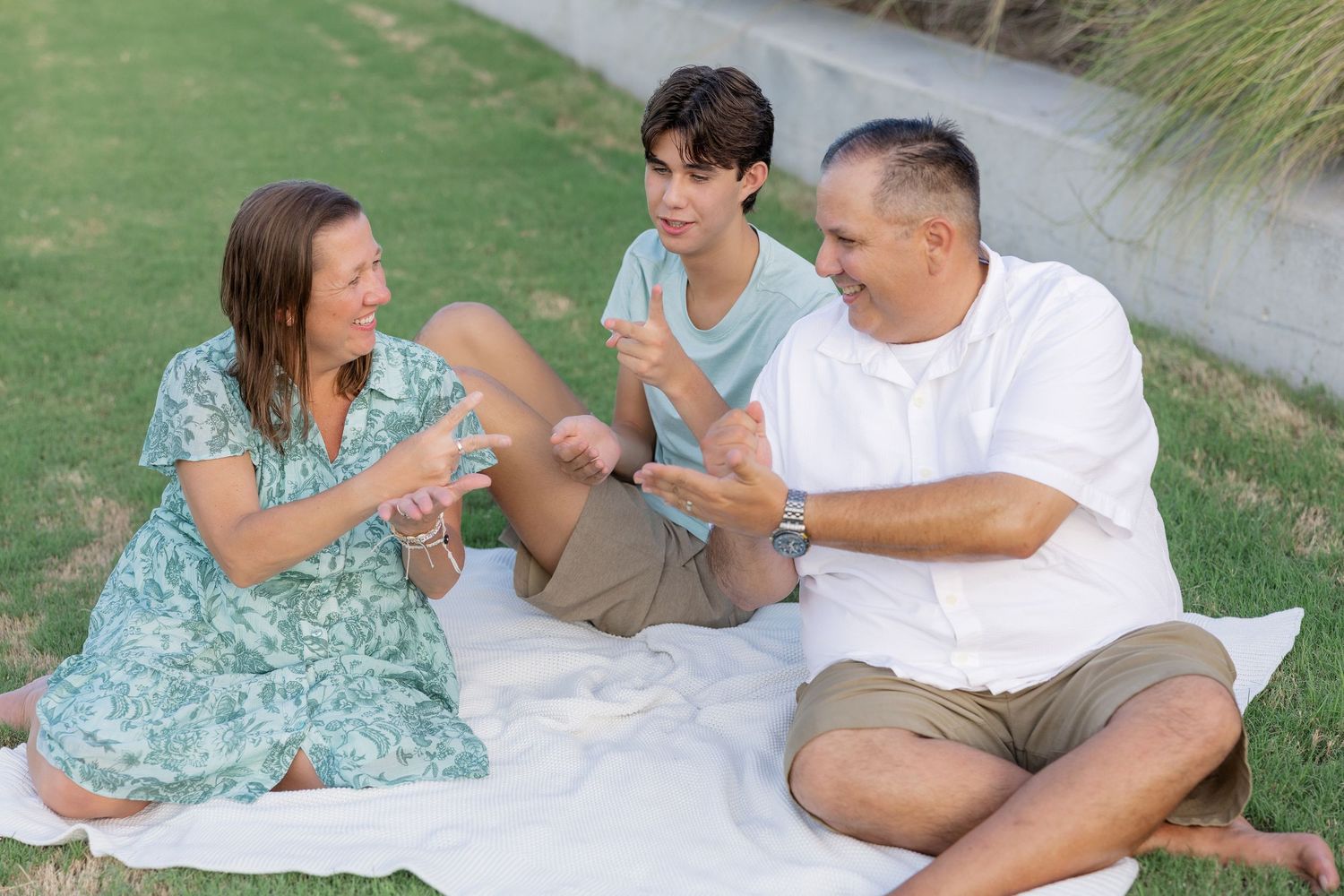 Family of three enjoying a picnic on green grass, sitting on white blanket, talking and laughing together outdoors.