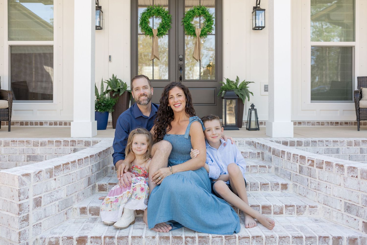 Family of four smiling on brick porch steps of white home with wreaths on door.