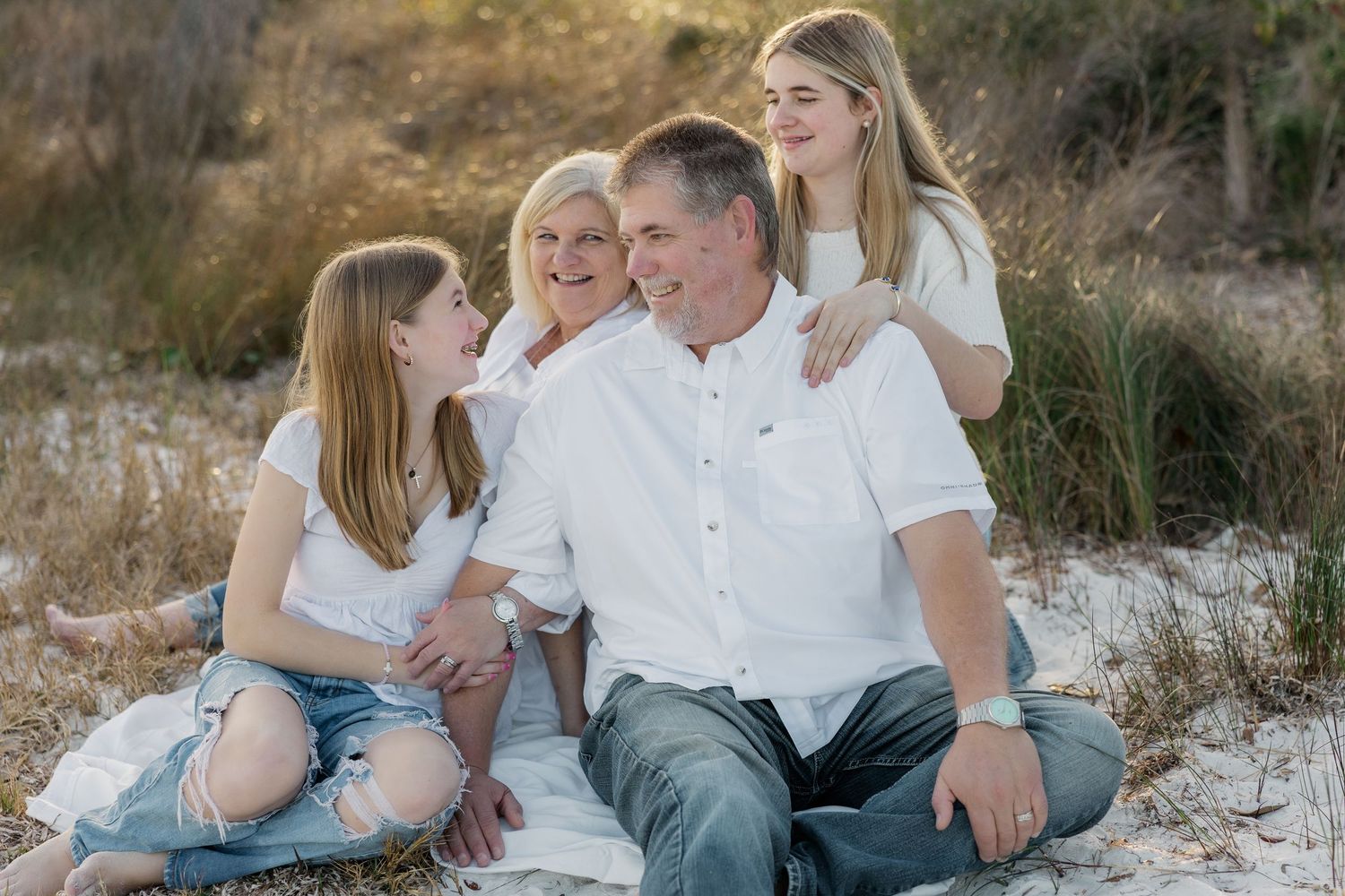 Family wearing coordinated white and blue outfits sits together on white sand beach during sunset photo session.