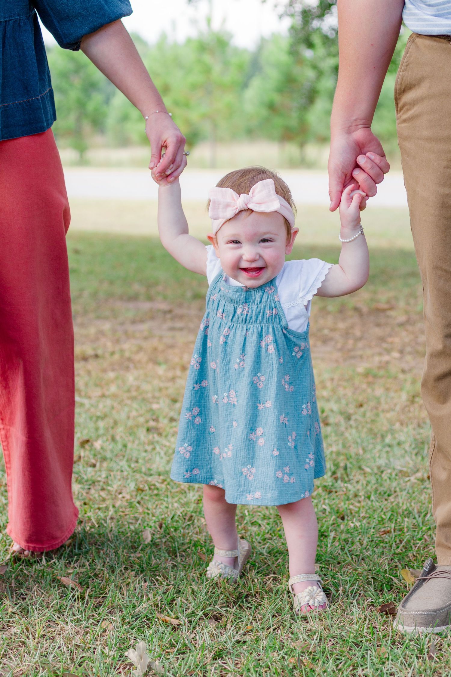 A baby in a light blue dress smiles while being supported by adults holding their hands on a grassy lawn.