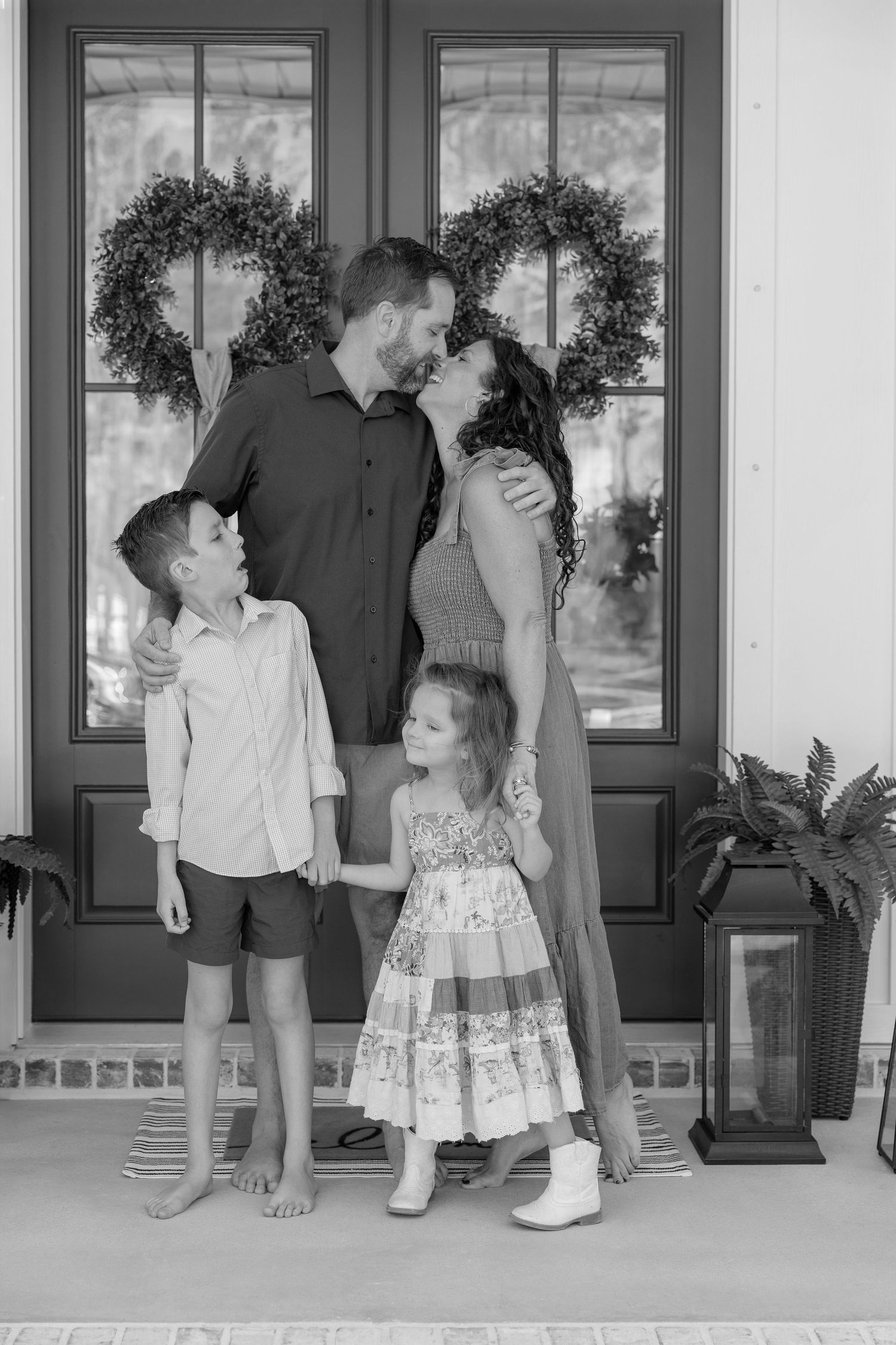 Black and white family portrait of parents kissing on porch with two young children standing beside them.