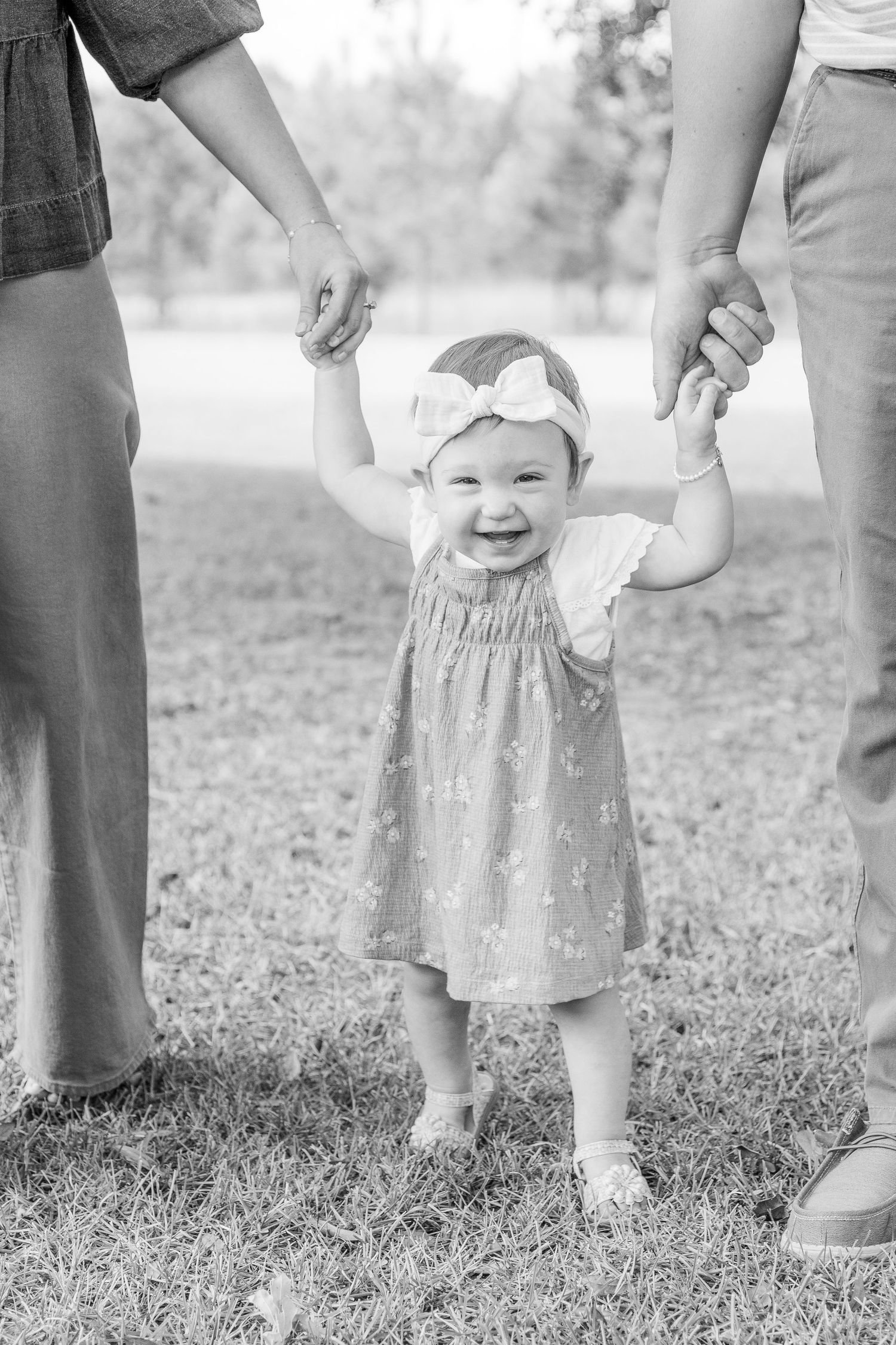 A small child in a dress takes first steps while holding hands with adults in an outdoor setting.