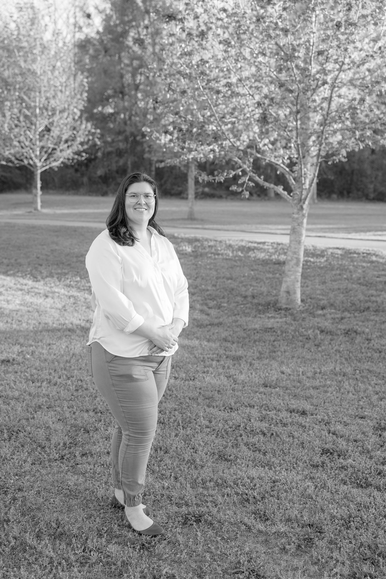 Black and white portrait of a smiling woman in white shirt and pants standing in a park with trees.