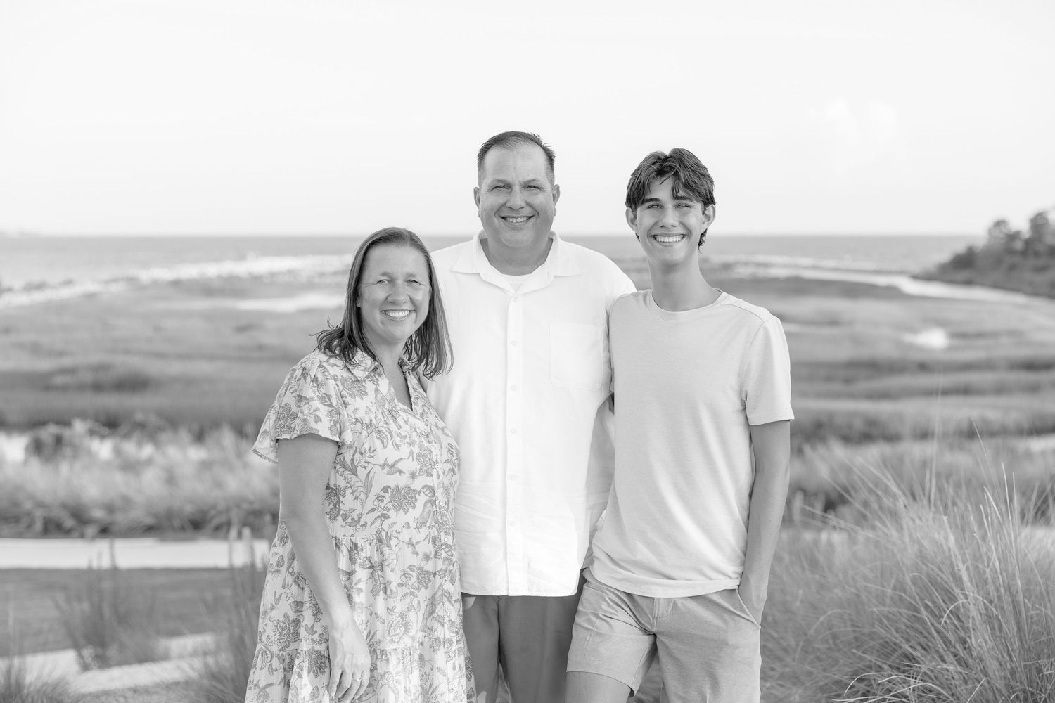 Black and white photo of a smiling family of three posing outdoors near a coastal marsh landscape.