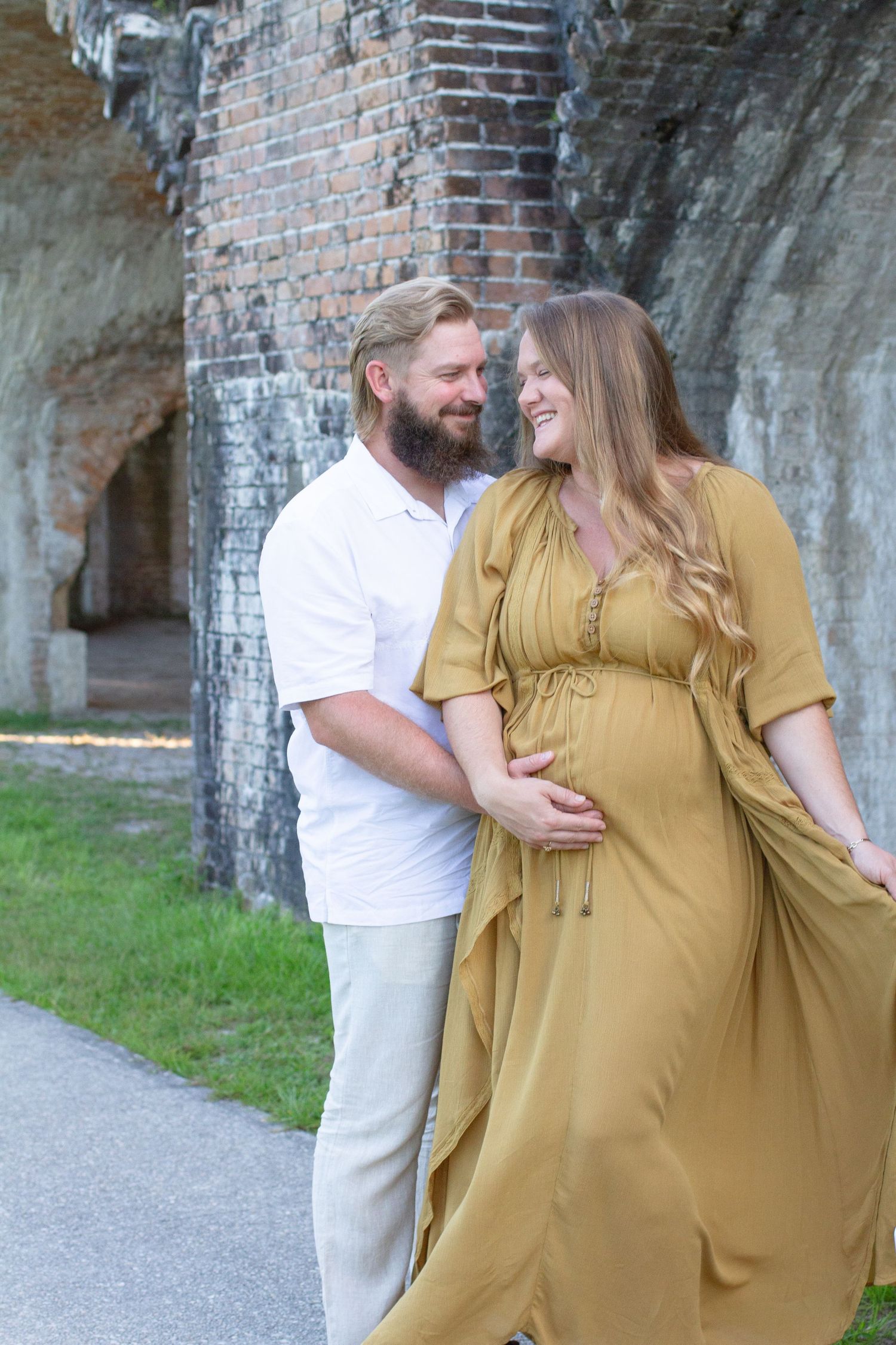 A couple shares a romantic moment together in front of a rustic brick wall backdrop on a paved path.