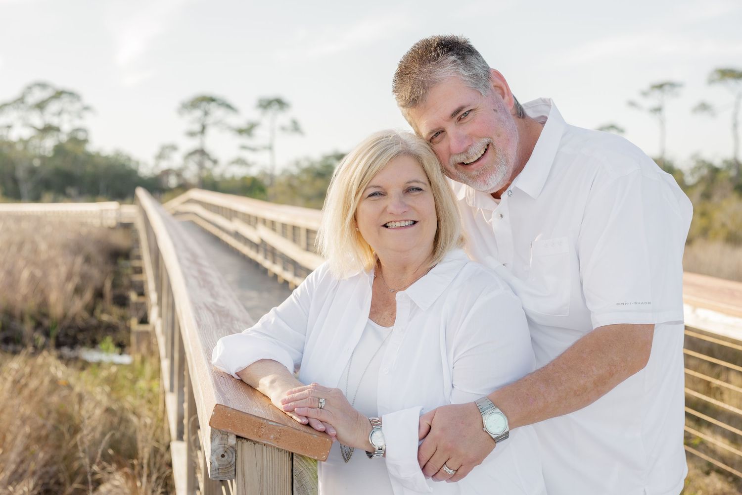 A couple wearing white clothing embraces while standing on a wooden boardwalk near the beach at sunset.