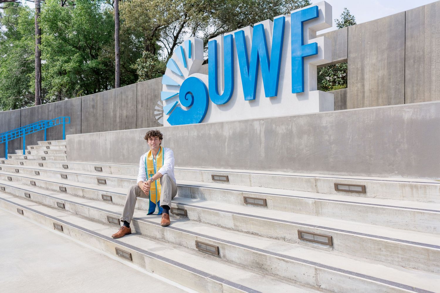 Graduate in regalia sits on concrete steps before a large blue UWF sign at the University of West Florida.