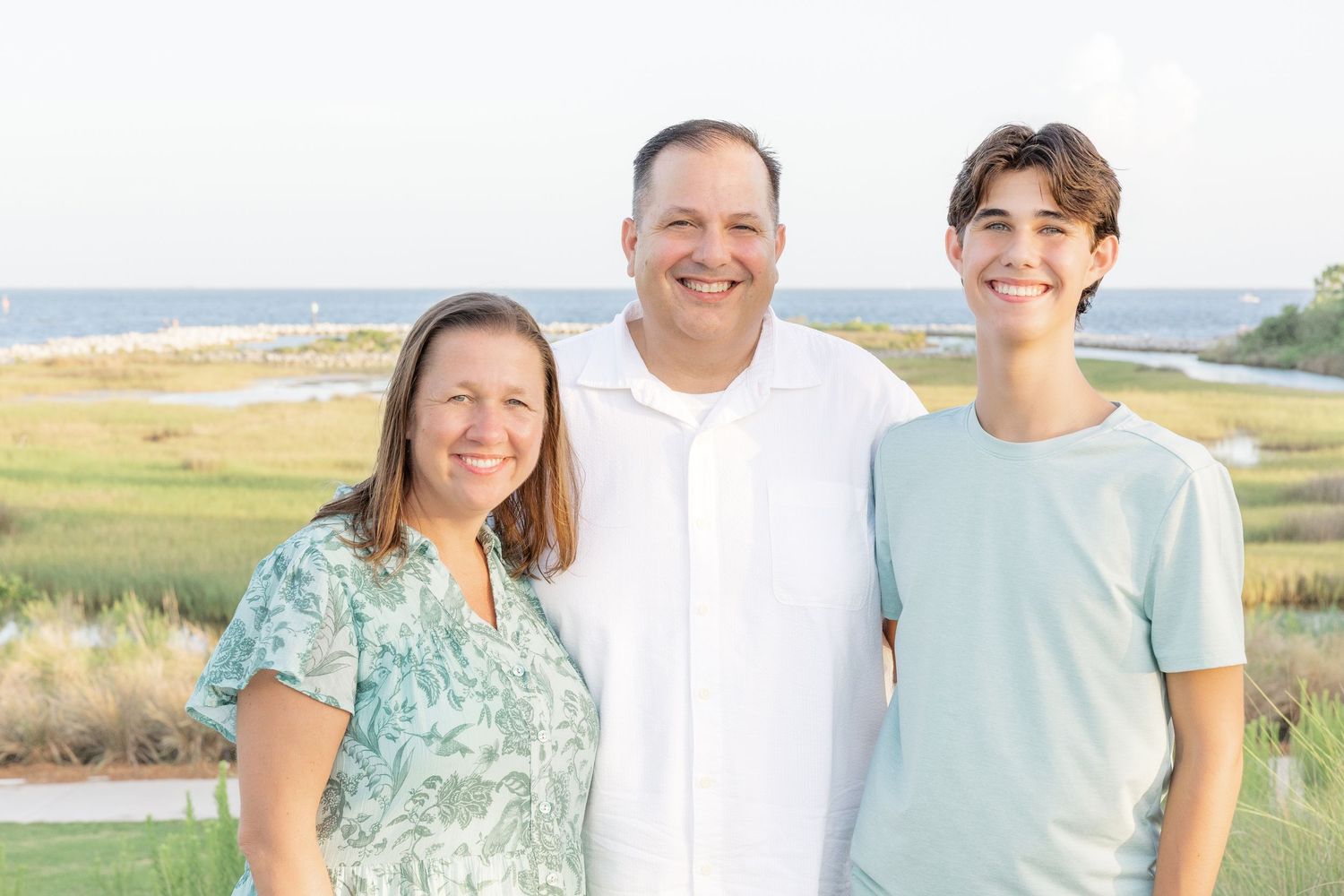 Family of three smiling outdoors near coastal marsh with ocean views on a sunny day.